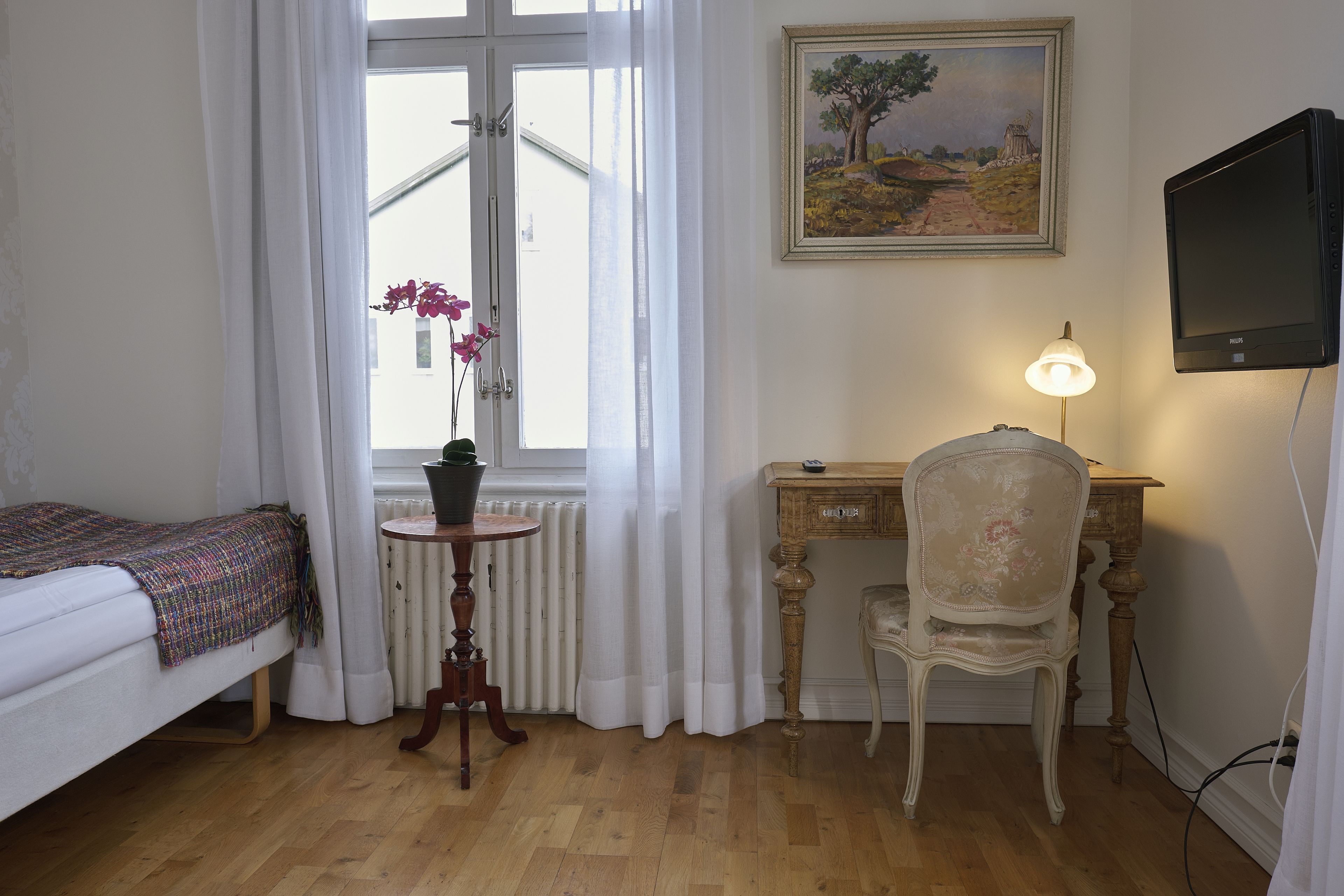 A bedroom with a bed, a small table with a potted plant, a desk with a chair, a painting on the wall, and a television.