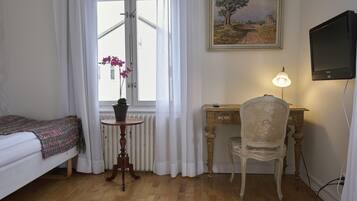 A bedroom with a bed, a small table with a potted plant, a desk with a chair, a painting on the wall, and a television.