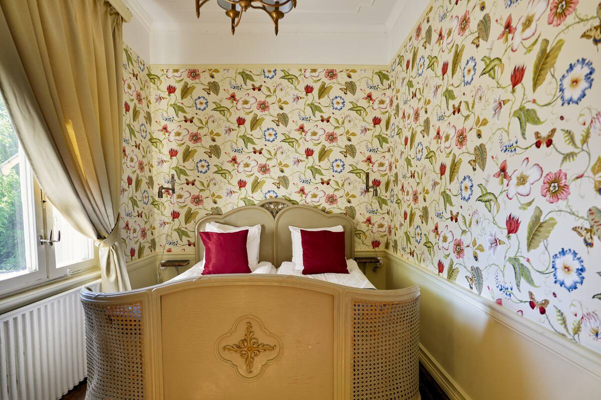 A bedroom with a bed, floral wallpaper, and a window with curtains.