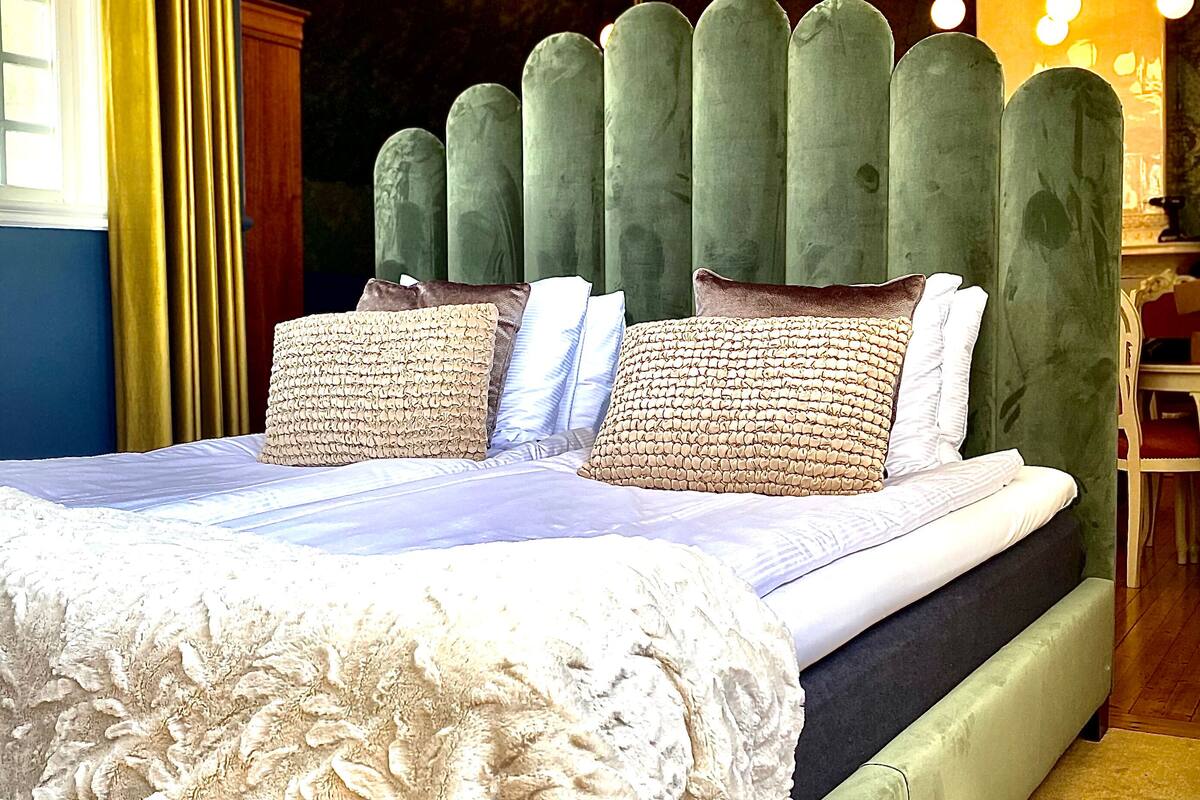A bedroom with a large bed, green upholstered headboard, multiple pillows, and a decorative ceiling light fixture.