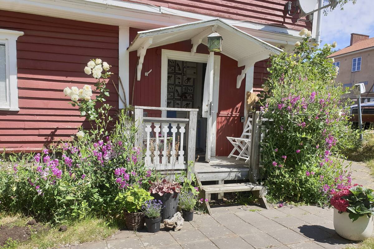 A red house with a white porch, a white door, and a white railing. There are potted plants and flowers around the porch.