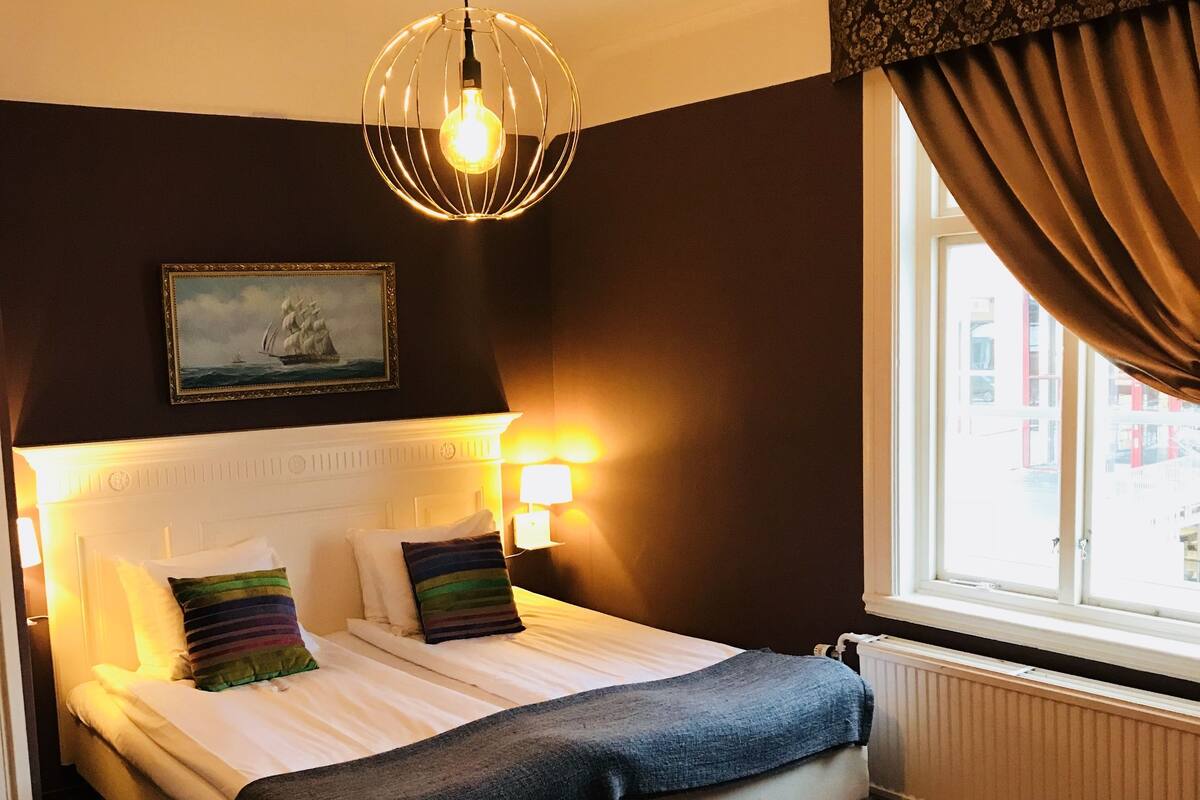 A neatly made bed with a headboard, a bedside lamp, and a painting of a ship on the wall.