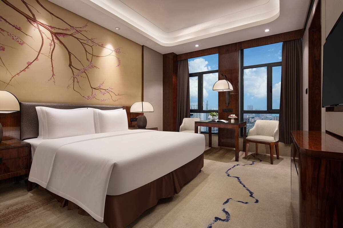 Une chambre d’hôtel avec un grand lit, un bureau, deux chaises, une télévision et une vue sur la ville grâce à de grandes fenêtres.