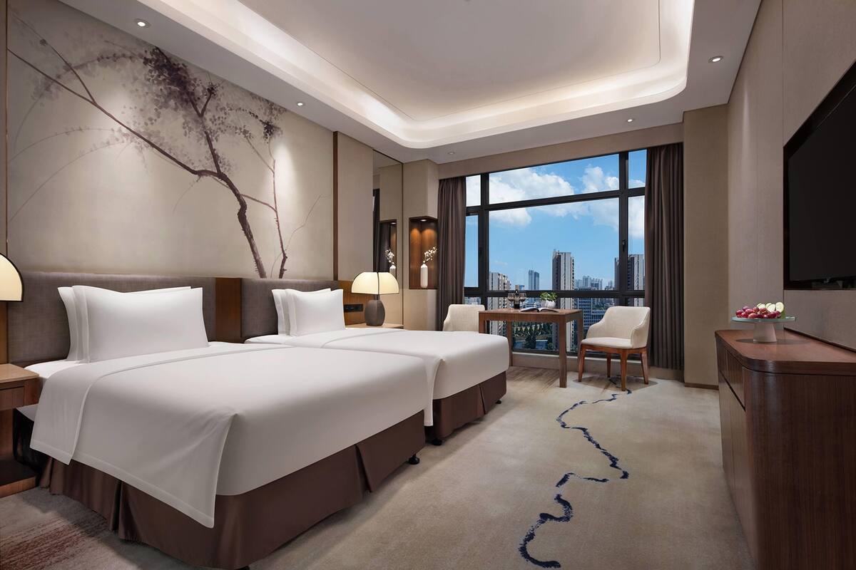 Une chambre d’hôtel avec deux lits, un bureau, une chaise, une télévision et une grande fenêtre donnant sur la ville.