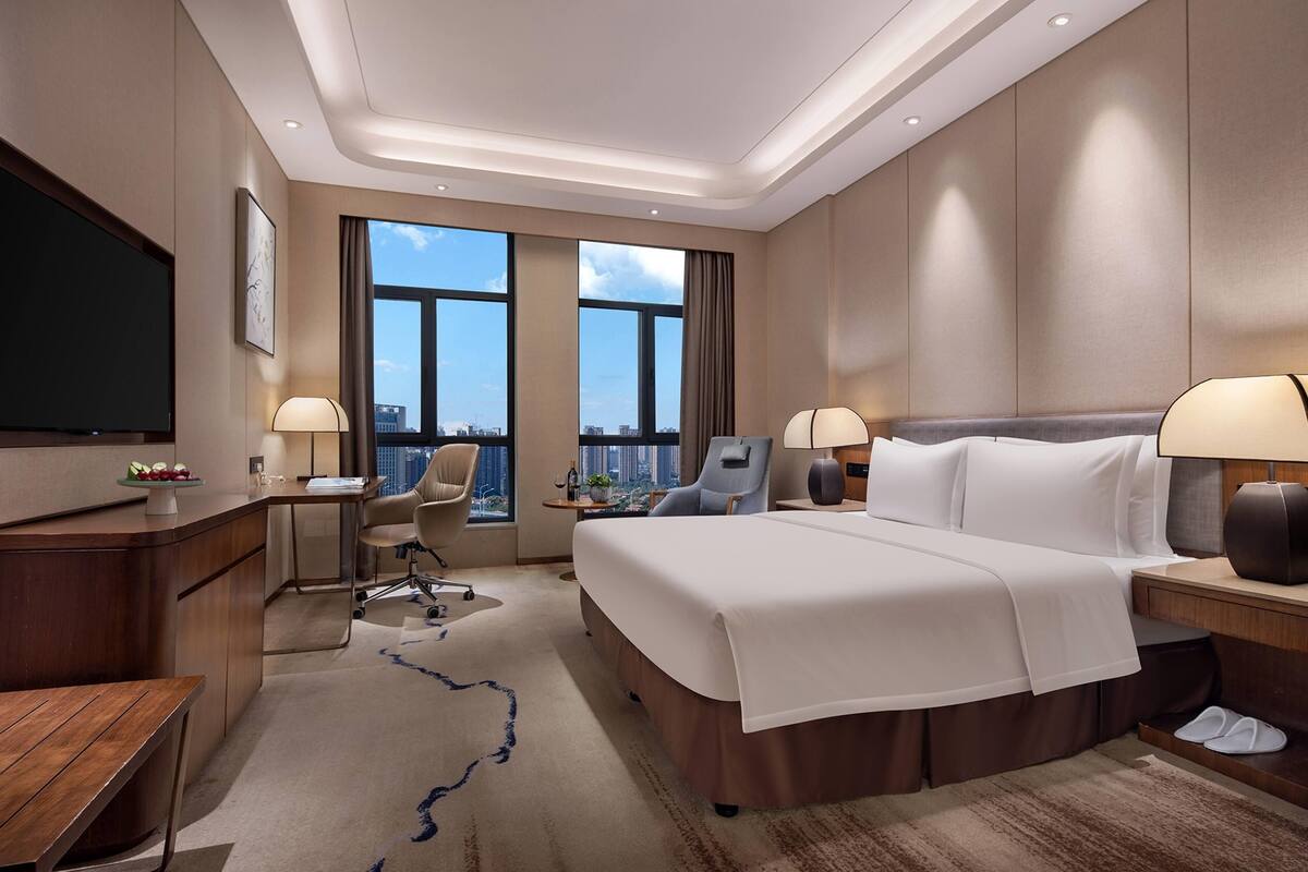 Une chambre d’hôtel dotée d’un grand lit, d’un bureau, d’une chaise, d’une télévision et offrant une vue sur la ville grâce à de grandes fenêtres.