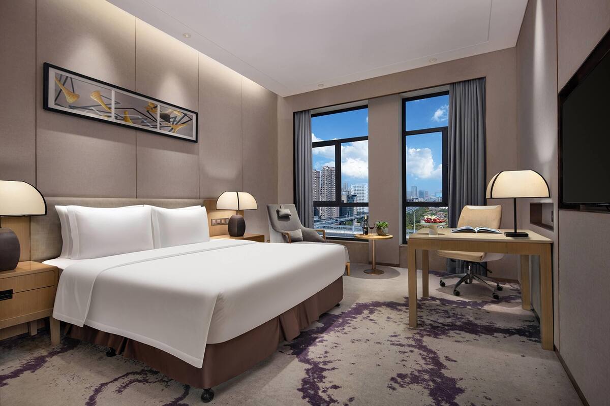 Une chambre d’hôtel avec un grand lit, deux tables de chevet avec des lampes, un bureau avec une chaise, une vue sur la ville et une grande fenêtre avec des rideaux.