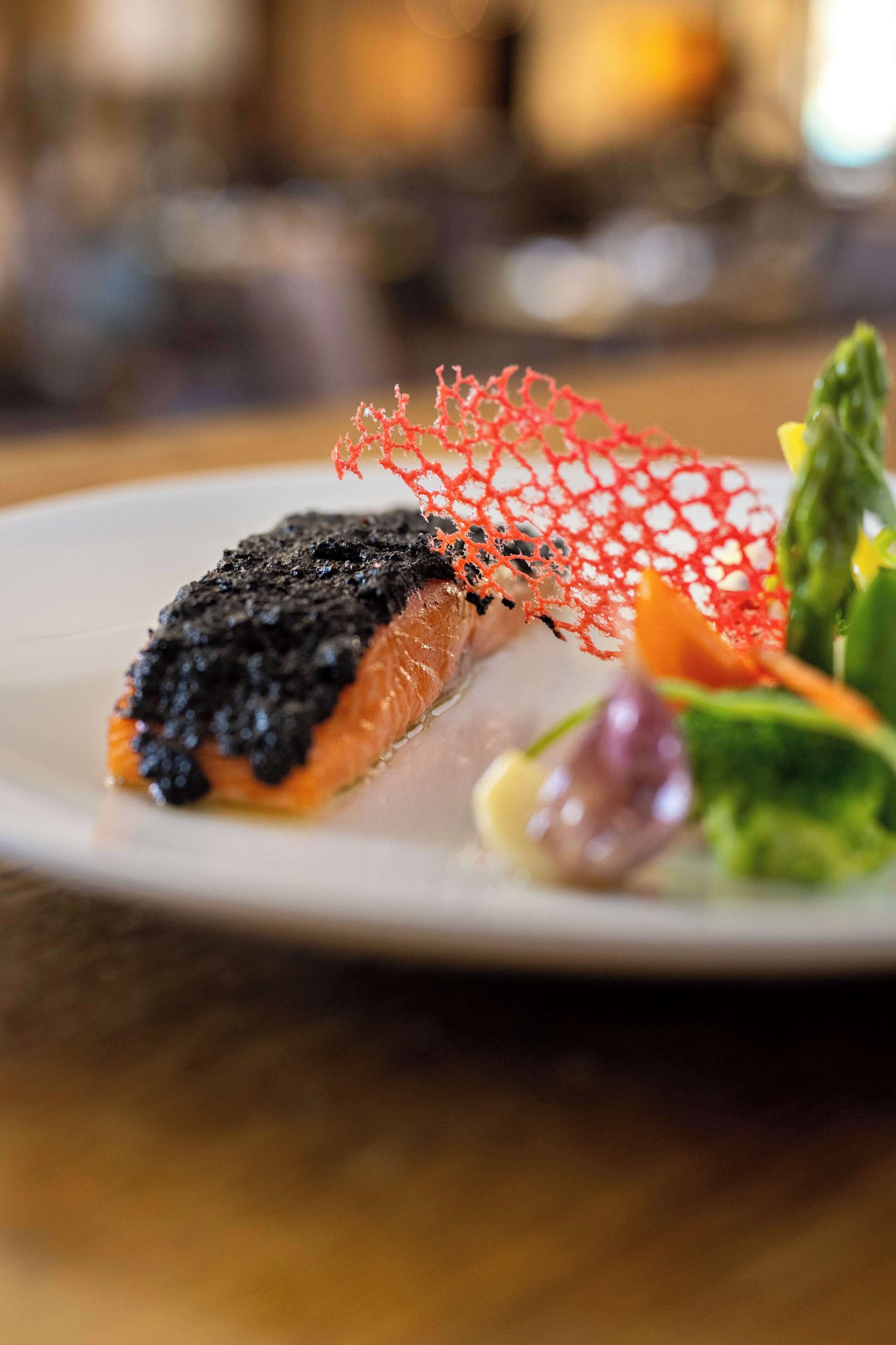 Un plato con un trozo de salmón cubierto de caviar negro y un adorno comestible rojo.
