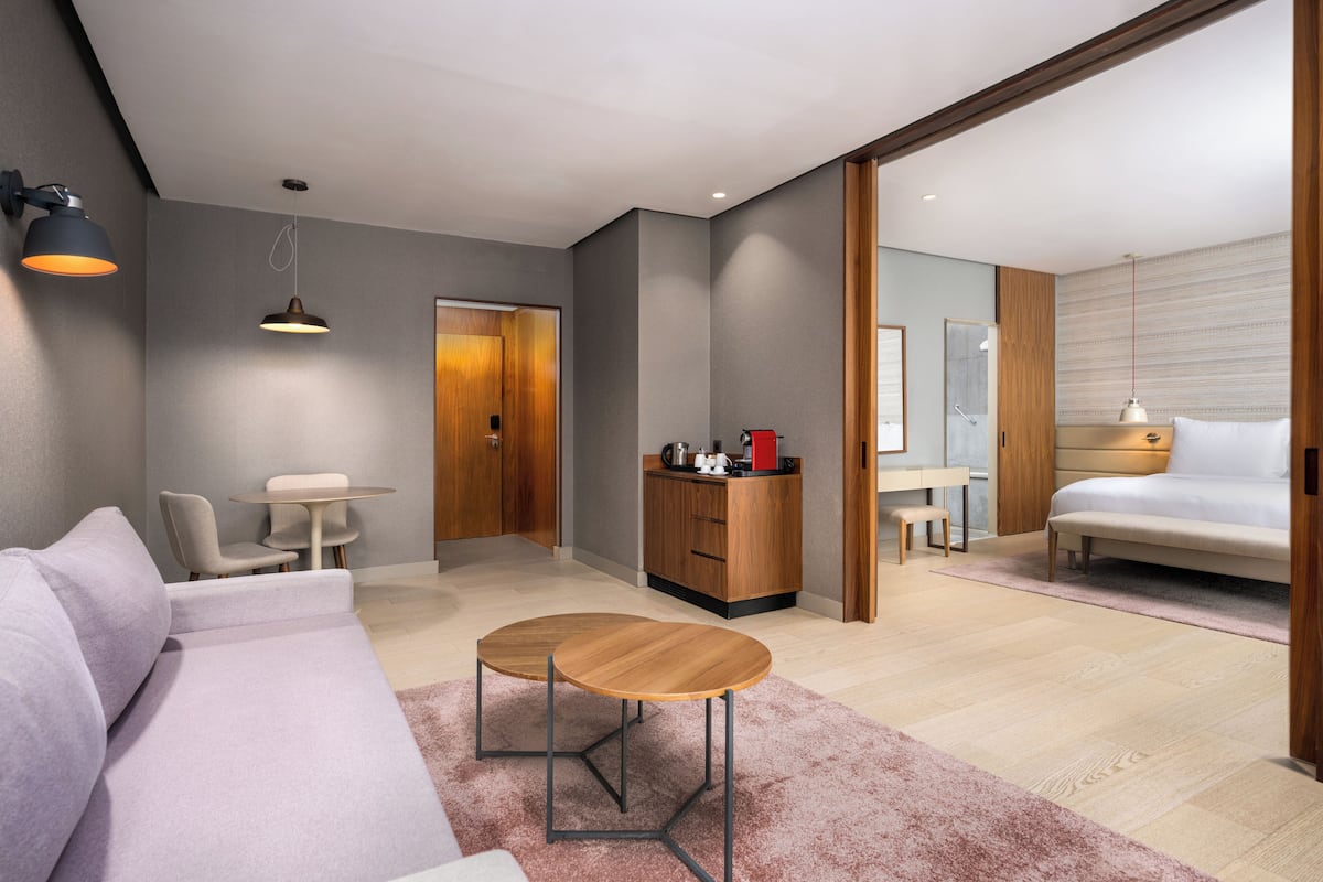 Une chambre d’hôtel moderne équipée d’un canapé, d’une petite table ronde, d’un lit, d’un bureau et d’une salle de bain visible à travers une porte ouverte.