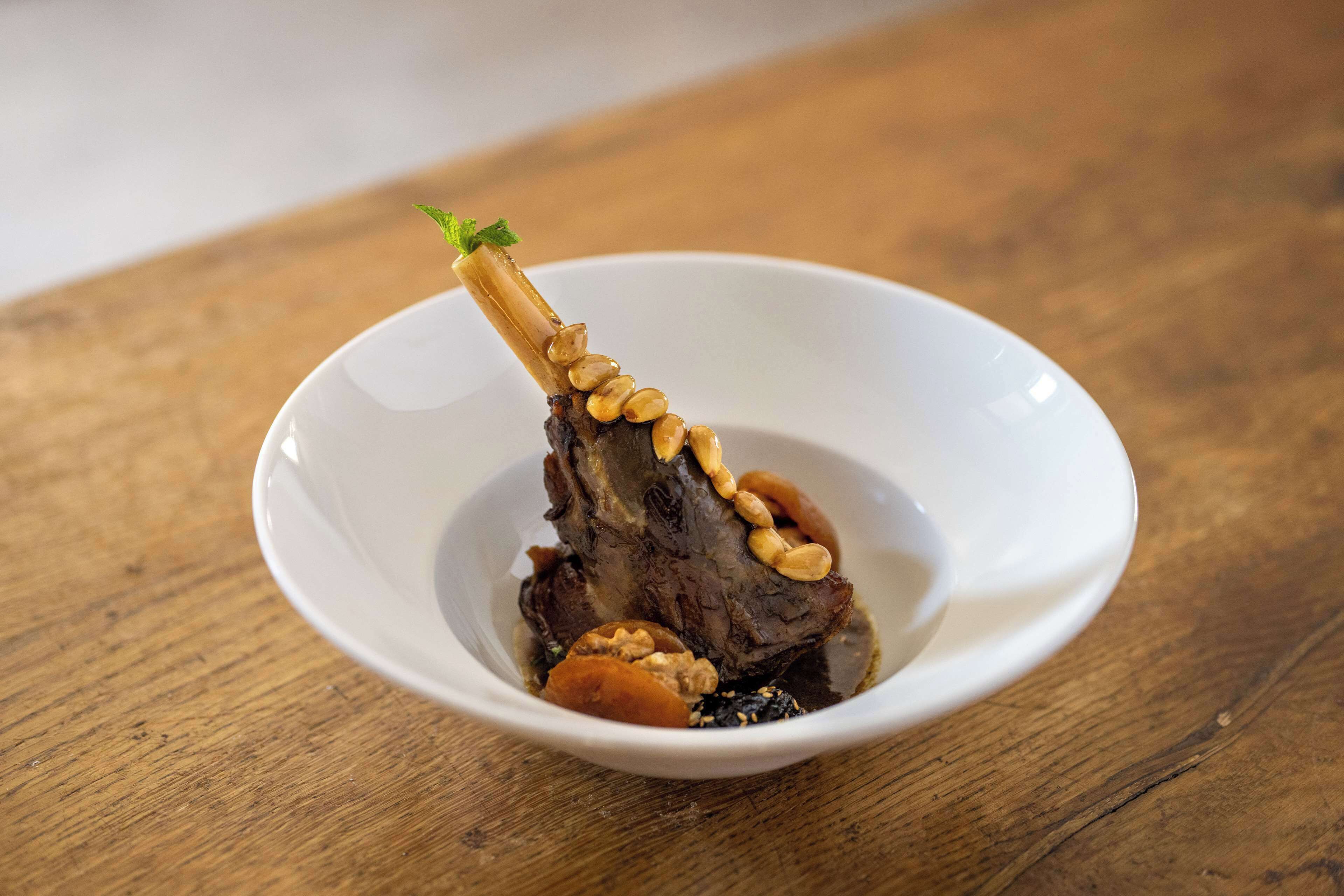 A white bowl containing a piece of meat with a bone, garnished with nuts and a sprig of herb, on a wooden surface.