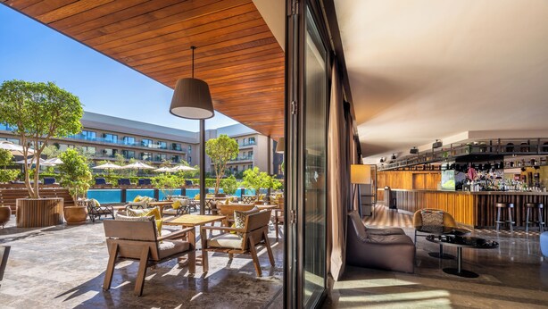 An outdoor seating area with tables and chairs, a bar with stools, and a view of a swimming pool and buildings in the background. - Radisson Blu Hotel, Marrakech Carré Eden (Marrakech)