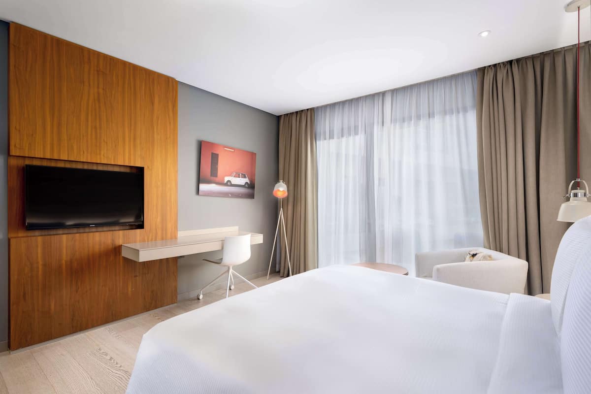 Une chambre d’hôtel moderne dotée d’un grand lit, d’un téléviseur à écran plat fixé sur un panneau en bois, d’un bureau avec une chaise, d’une petite table et d’une grande fenêtre avec des rideaux.