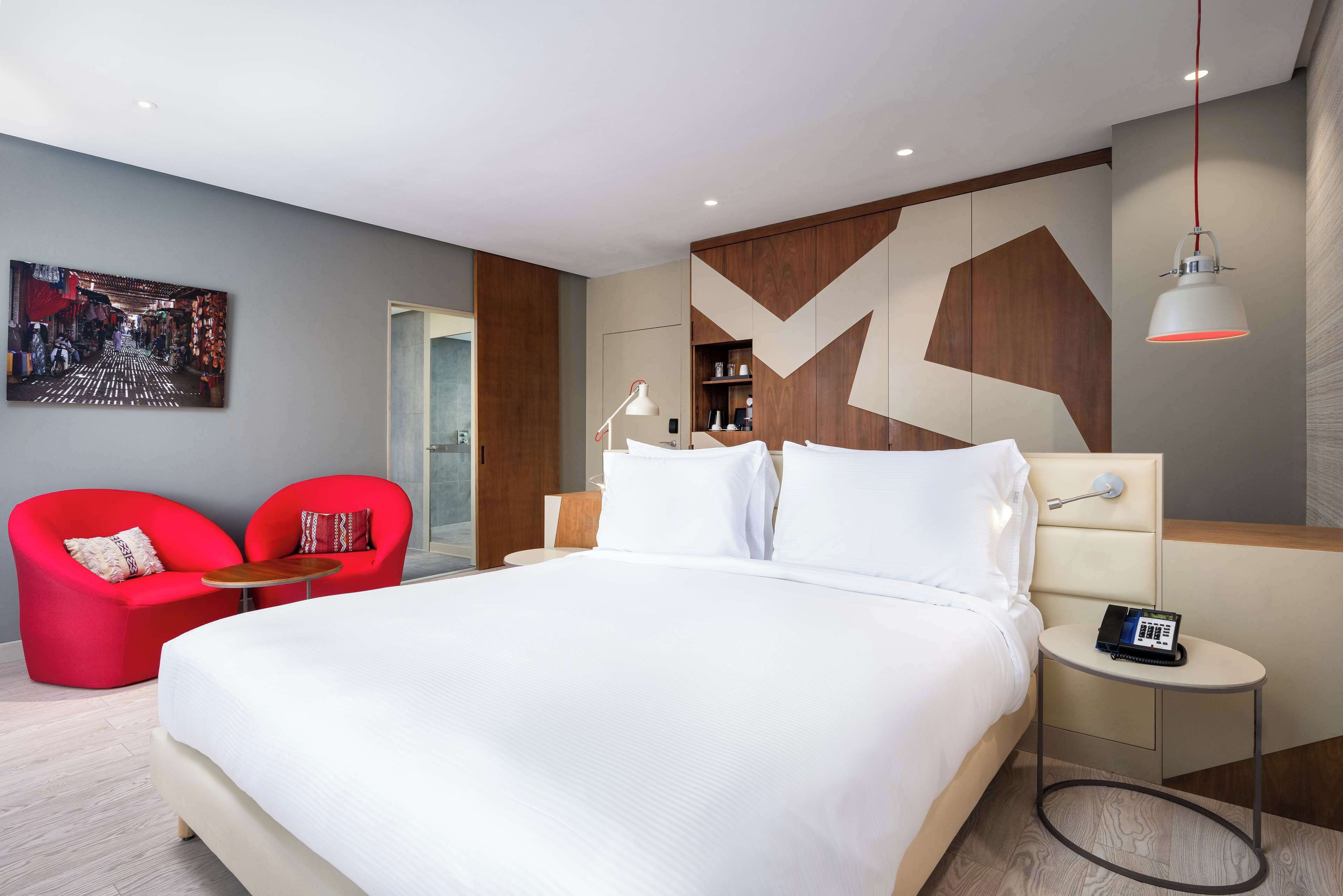 Una habitación de hotel moderna con una cama grande, un sillón rojo, una mesita redonda y una lámpara de noche.