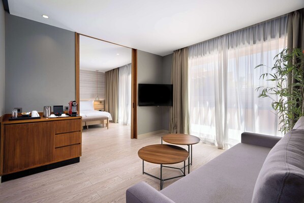 A modern hotel room with a wooden desk, two round tables, a sofa, a bed, and a TV.