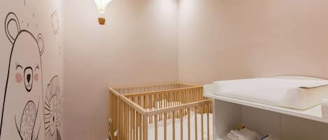 A baby room with a crib, a wall with a cute animal mural, a hanging light fixture, and a shelf with folded towels.