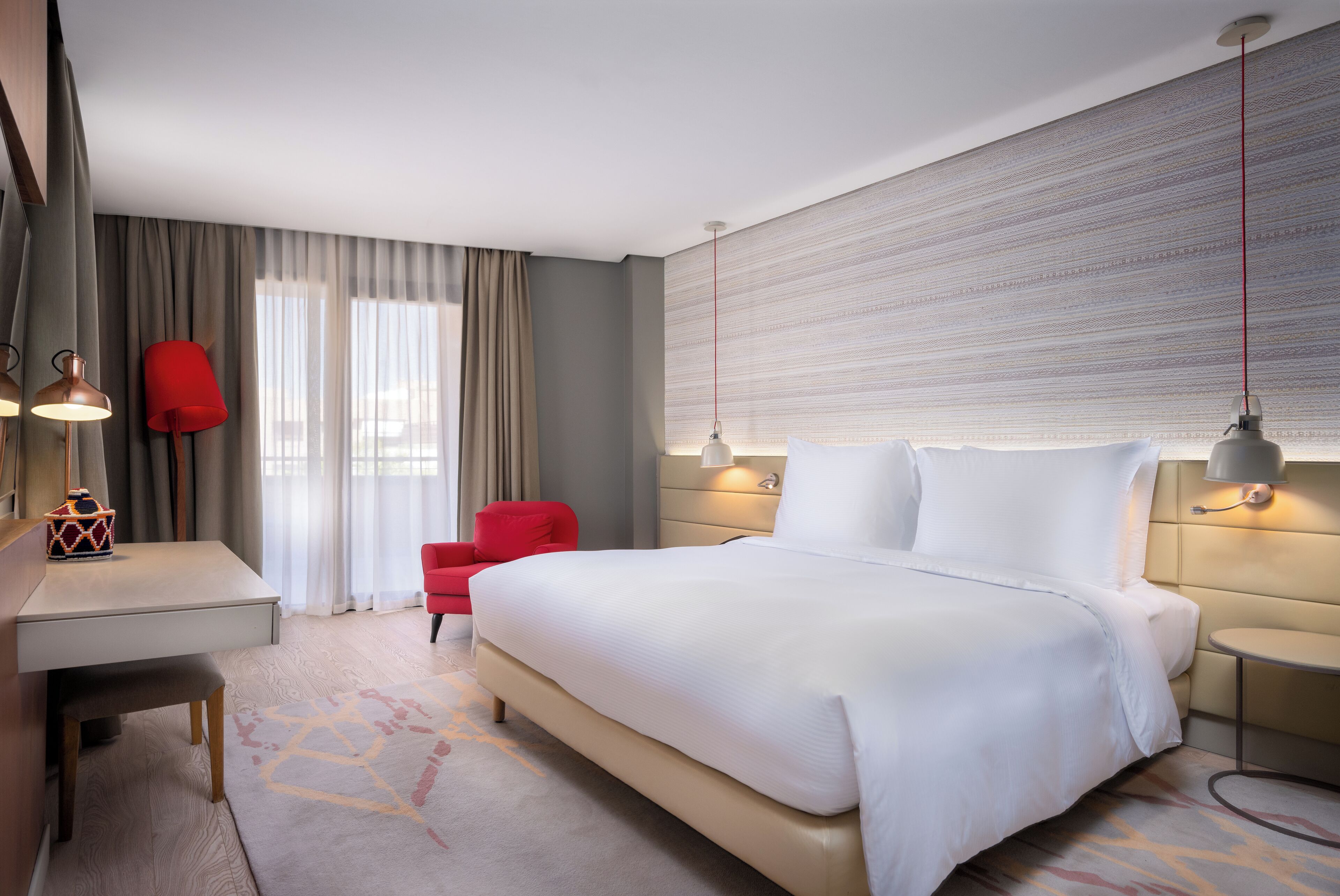 A modern hotel room with a large bed, a desk, a red chair, and a wall-mounted lamp.