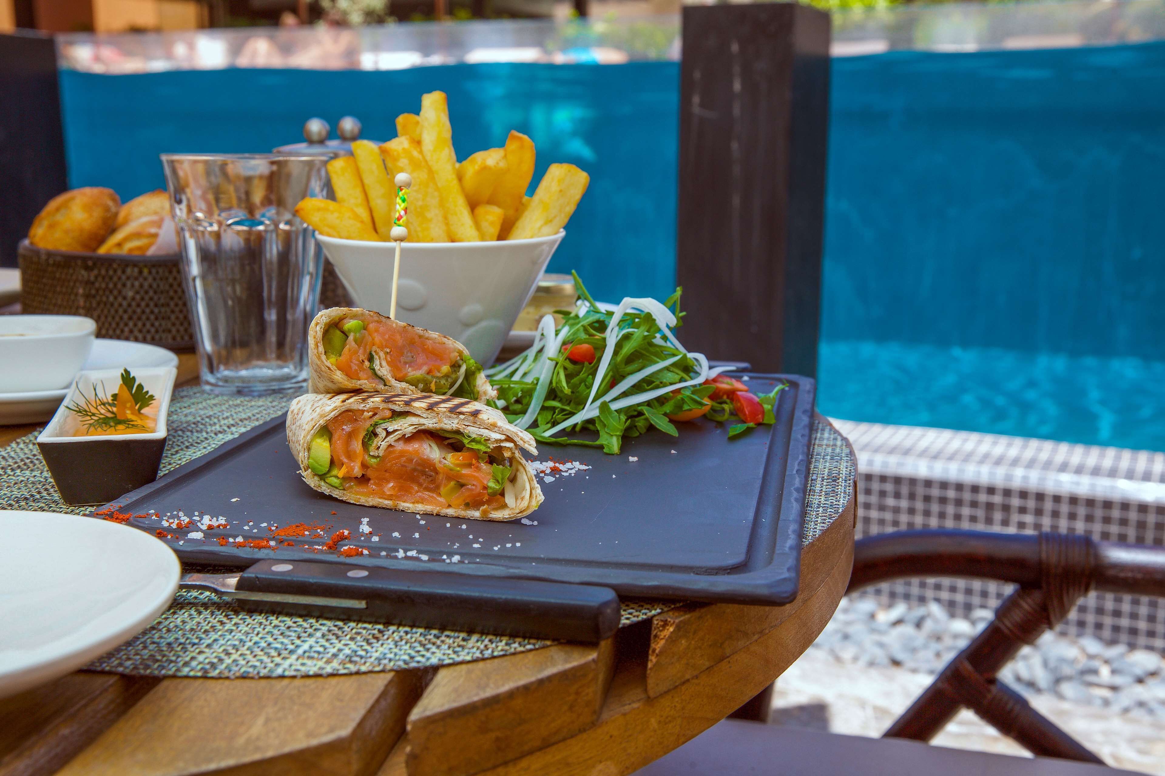 Comida con un wrap, papas fritas y ensalada sobre una mesa cerca de una piscina.