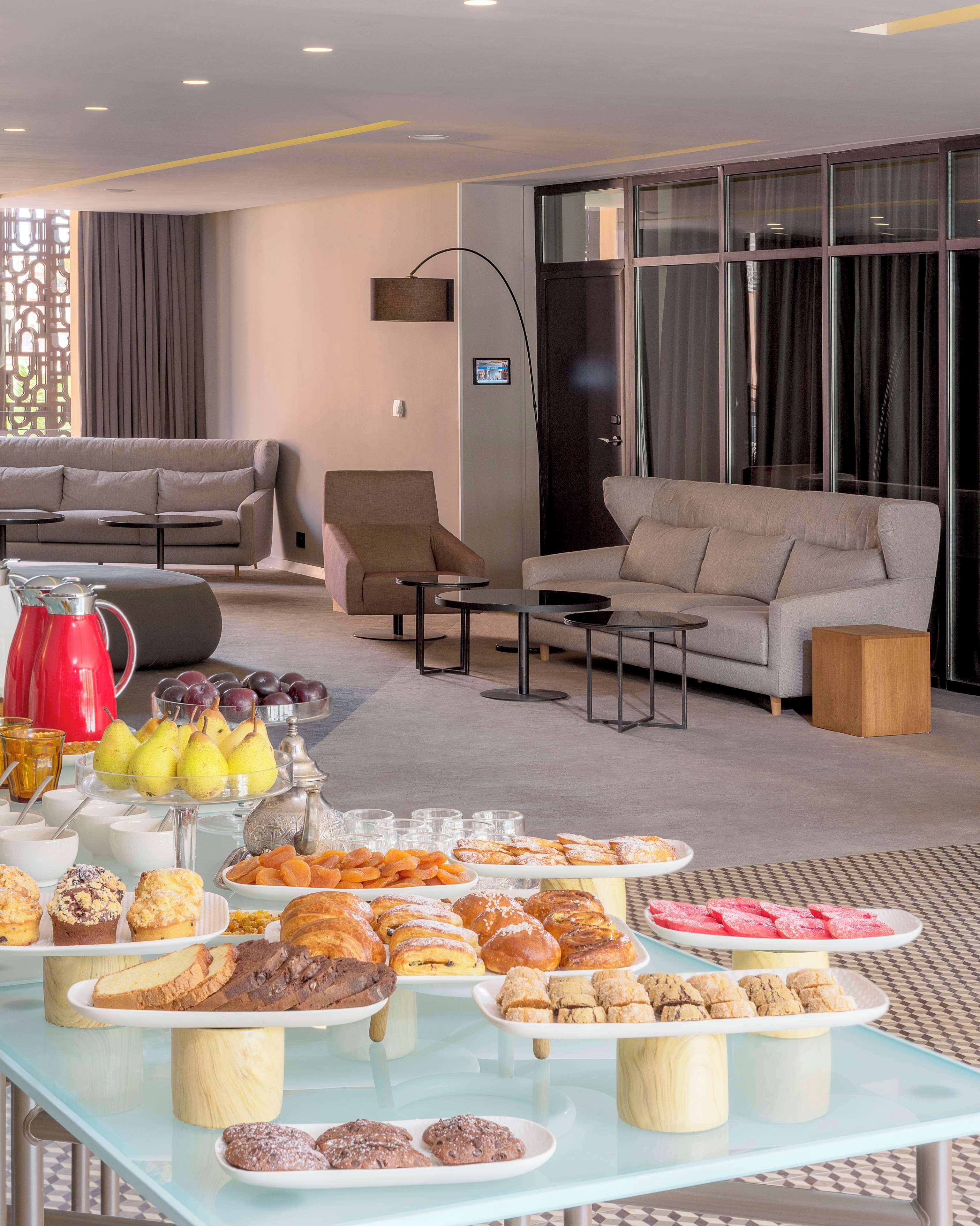Una sala moderna con un sofá, sillones y una mesa de centro. En primer plano, un aparador con diversos pasteles y frutas.