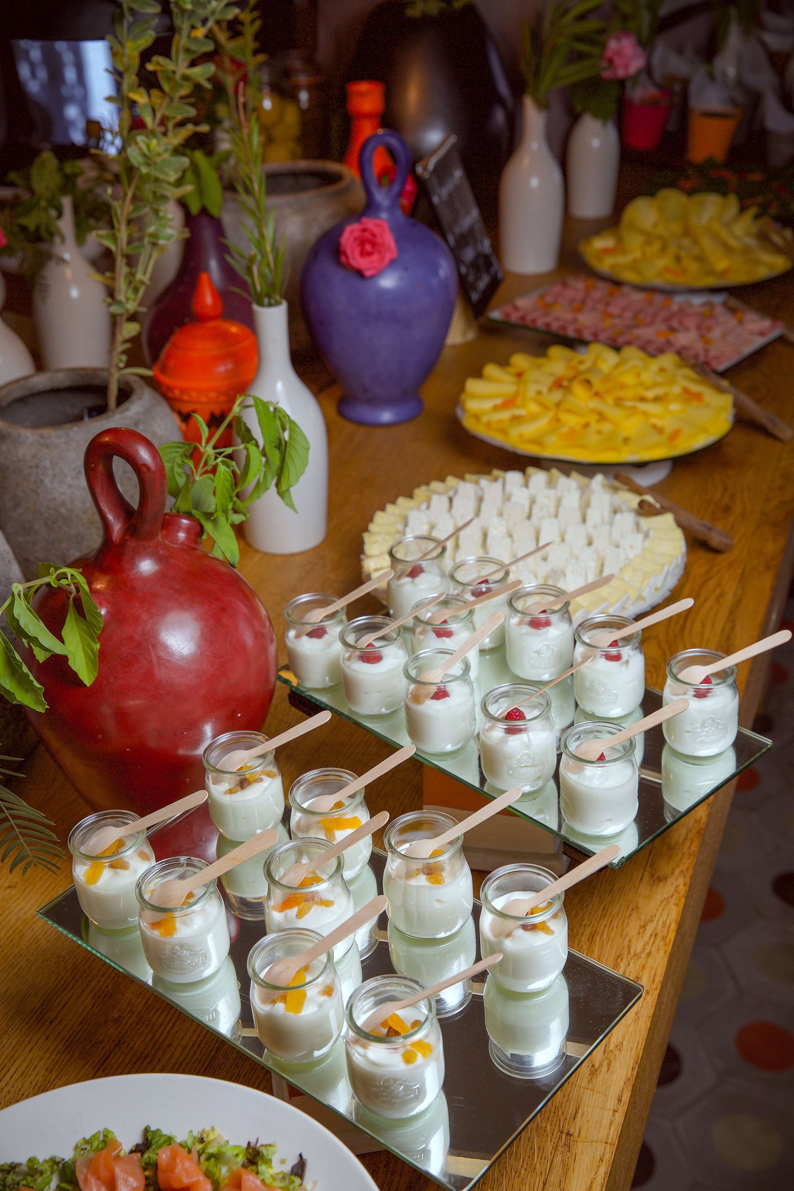 Una mesa de buffet con diversos alimentos, incluyendo una jarra roja, pequeños frascos de comida y un plato de fruta cortada.