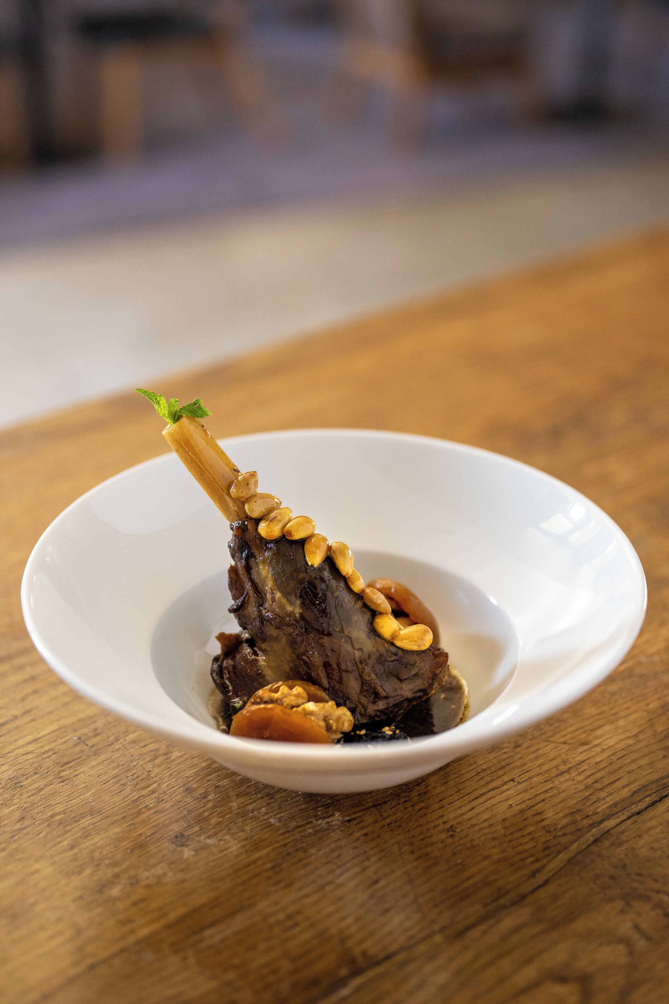 Un plato blanco con un trozo de carne adornado con nueces y una ramita de menta sobre una mesa de madera.