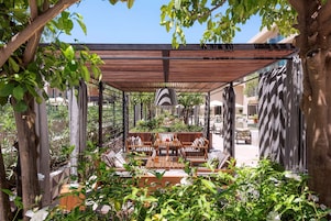 Un patio al aire libre con mobiliario de madera, una pérgola y abundante follaje.