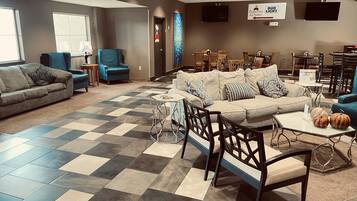 A spacious living area with a checkered floor, a grey sofa, a beige sofa, and a coffee table with pumpkins. There are blue chairs, wooden tables, and a wall-mounted TV.