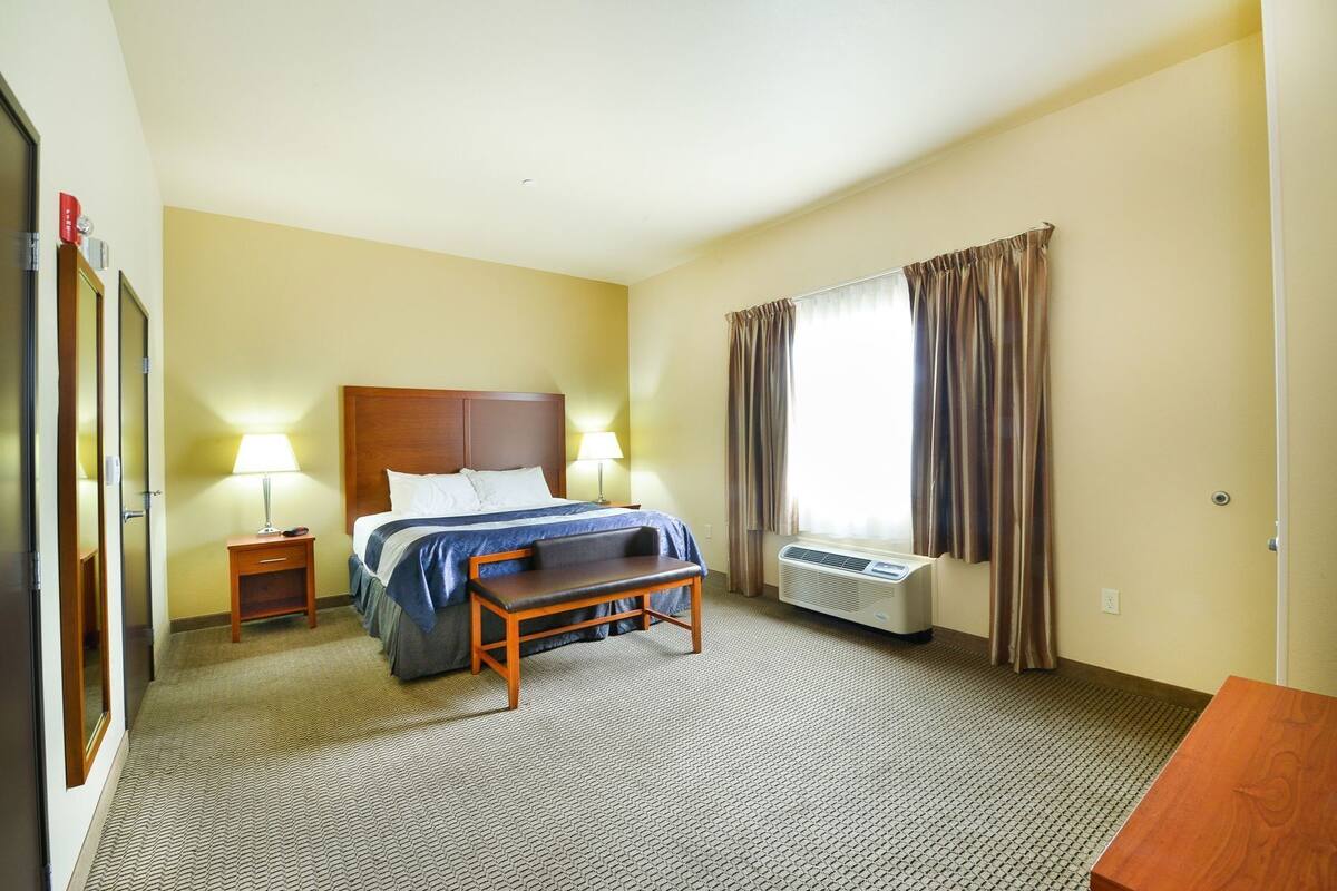 A hotel room with a bed, bedside lamps, a heater, a small table, and a window with curtains.
