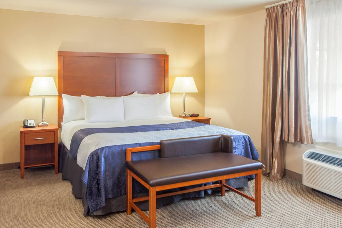 A hotel room with a large bed, two bedside tables with lamps, a bench, and a window with curtains.
