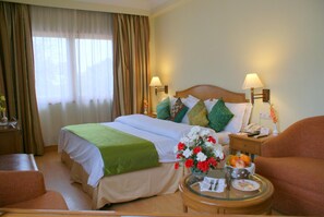 A hotel room with a bed, two bedside lamps, a nightstand with a phone, a small table with a vase of flowers and fruit, and a chair.