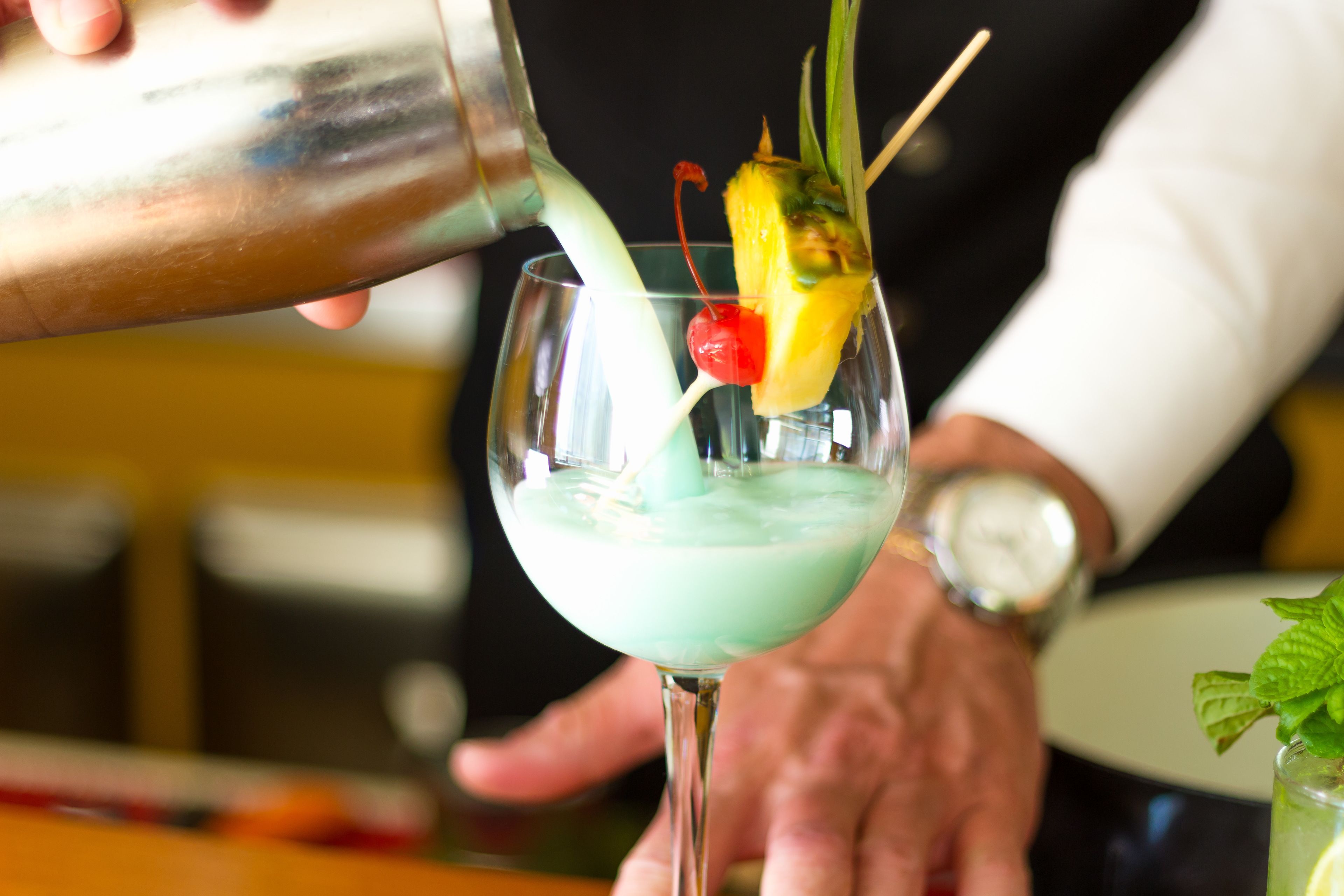 A bartender pouring a drink into a glass with a cherry and a slice of pineapple.
