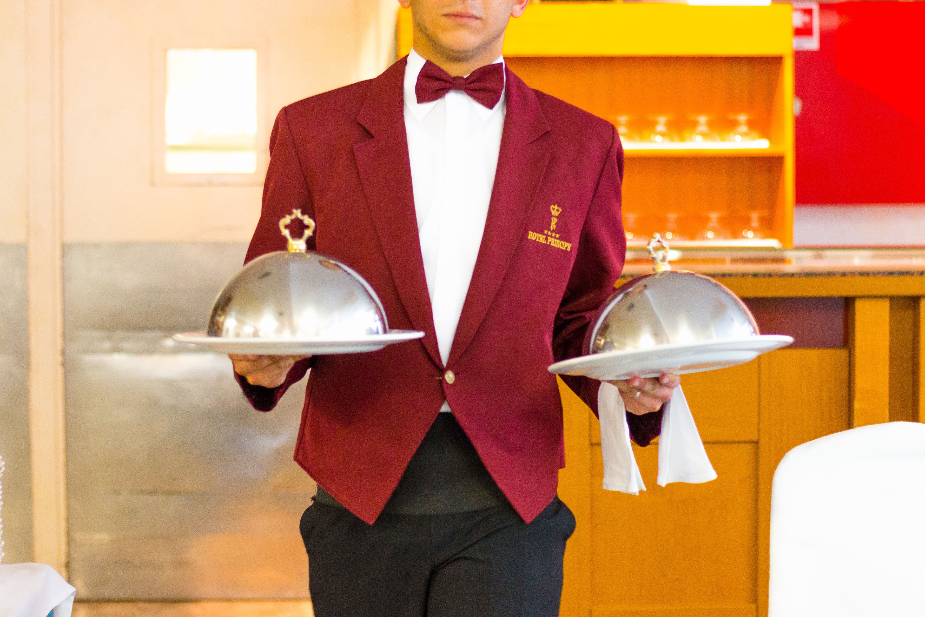 Un serveur vêtu d’un costume rouge, tenant deux plateaux en argent ornés d’un emblème doré, se tient dans une pièce où l’on aperçoit un comptoir en bois et un mur rouge.