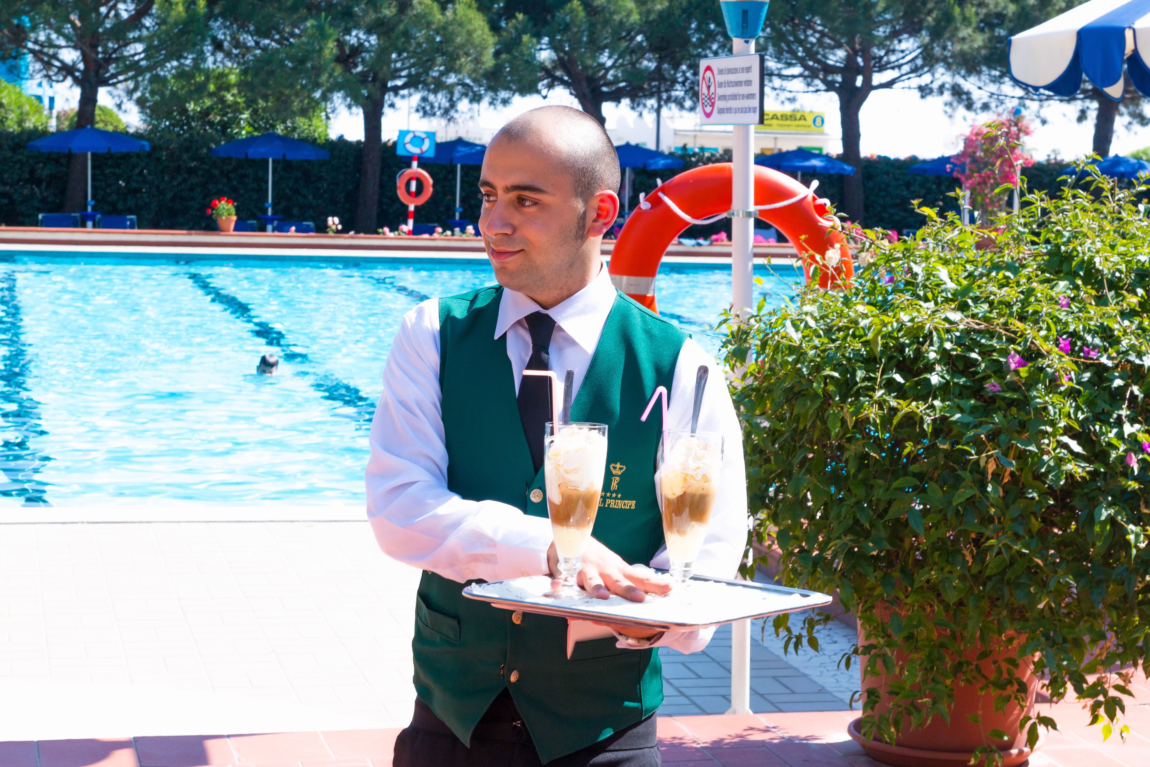 Escena junto a la piscina con un flotador salvavidas, plantas en macetas y una persona sosteniendo una bandeja con bebidas.