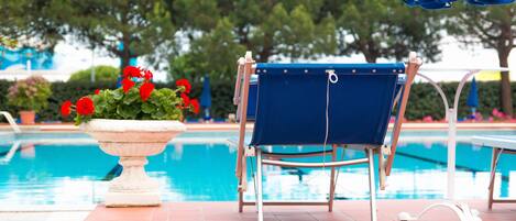A poolside area with a blue lounge chair, a white planter with red flowers, and a pool in the background.