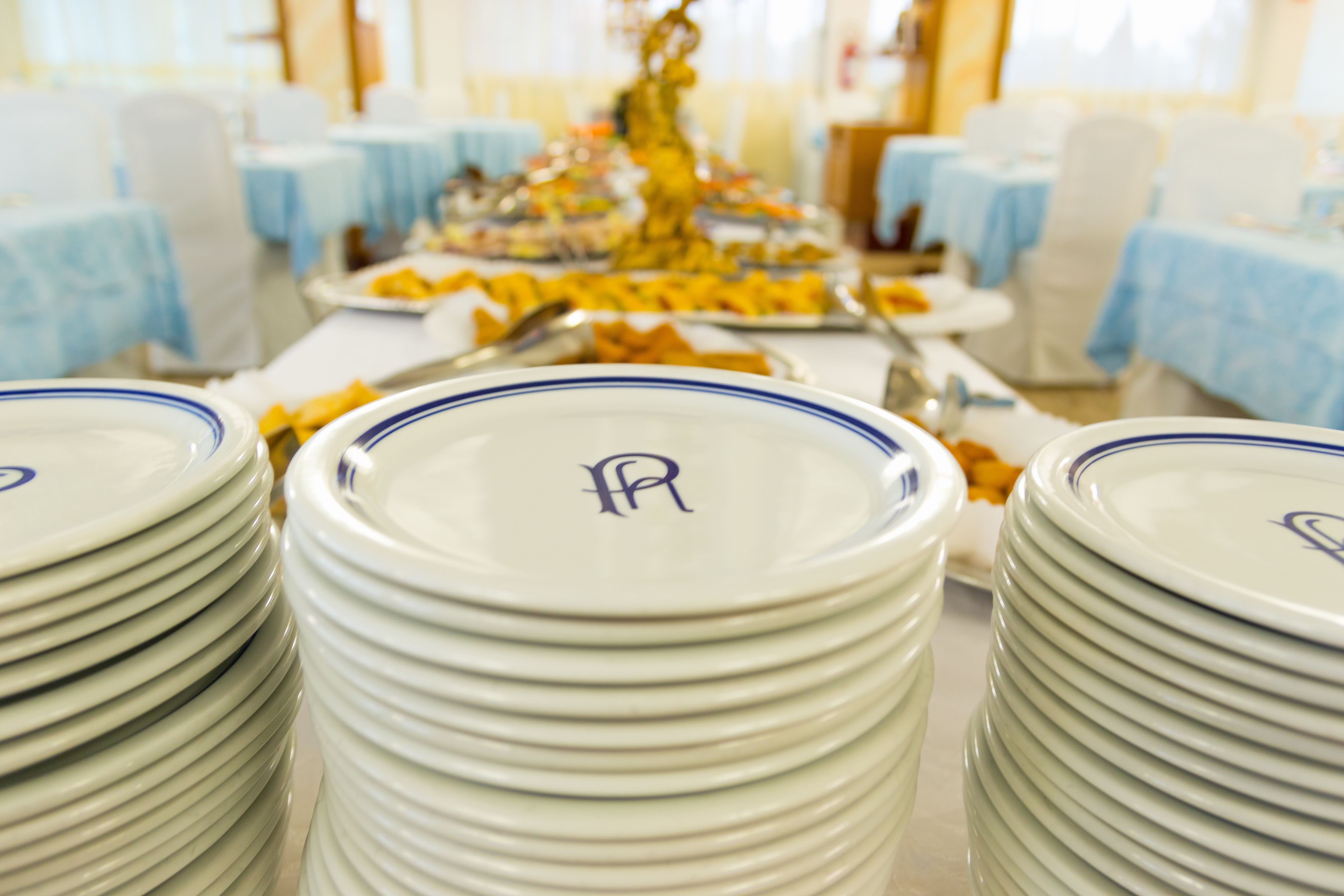 Stacks of white plates with blue rims and a monogram, arranged on a table in a room with blue tablecloths and white chairs.