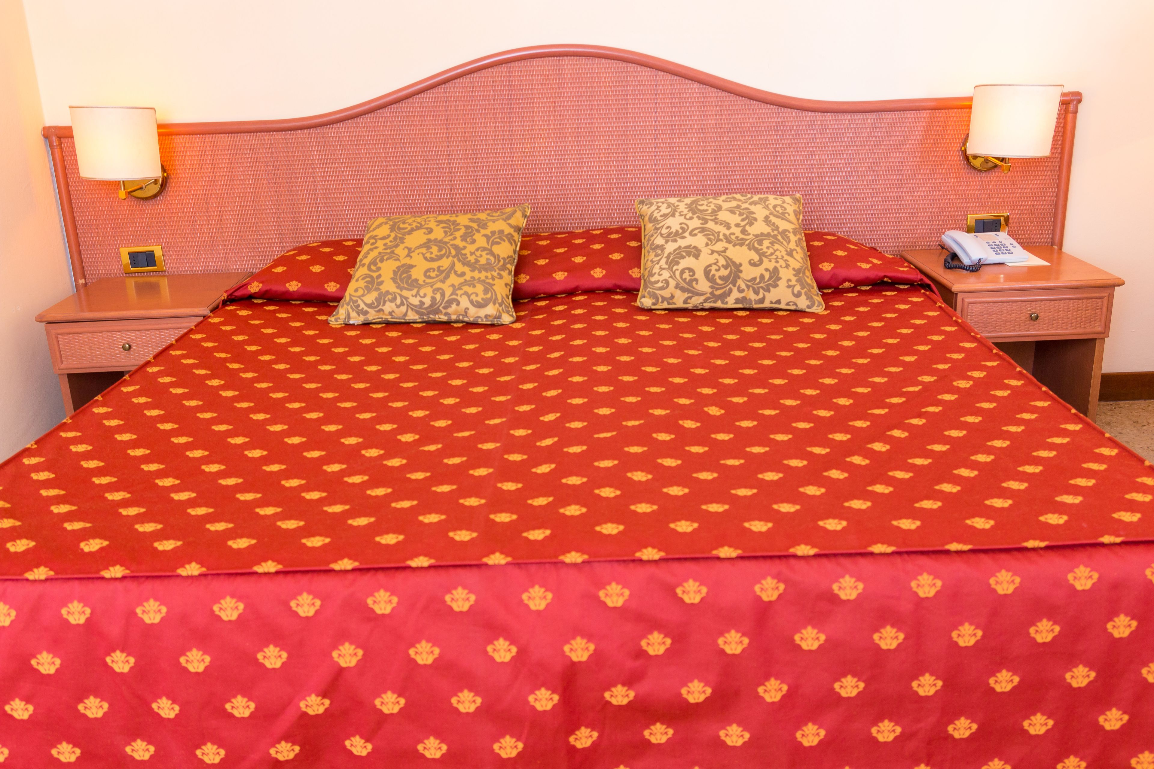 Una cama con una colcha roja estampada y dos almohadones decorativos. Se ven dos mesitas de noche, una de ellas con lámpara y teléfono.