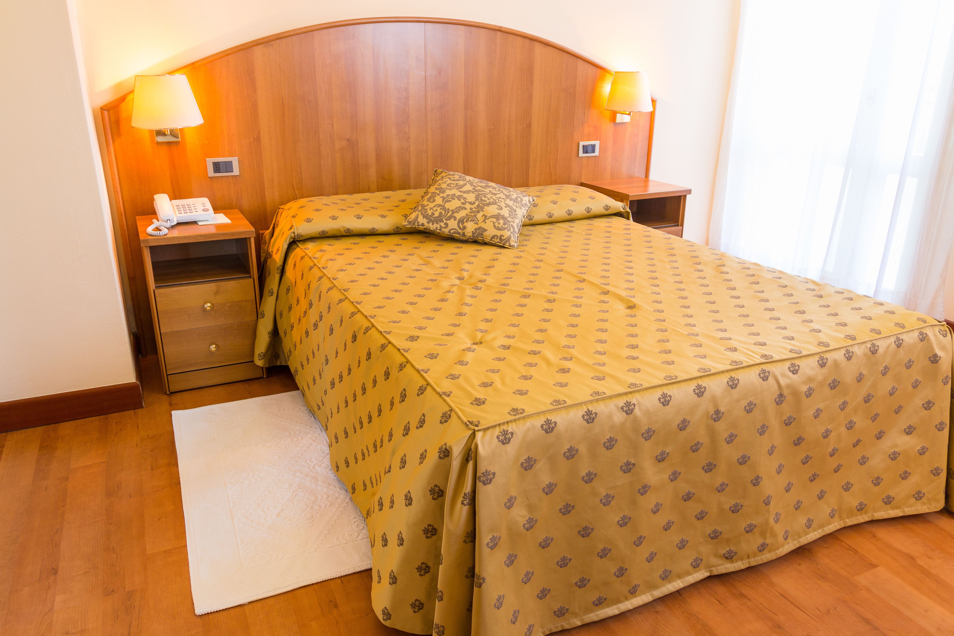 A hotel room with a wooden headboard, a bed with a patterned bedspread, two bedside lamps, a bedside phone, and a white rug.