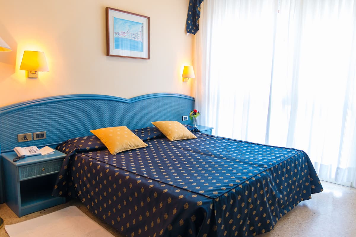 Ein Hotelzimmer mit blauem Kopfteil, zwei Nachttischlampen, einem Bett mit dunkel gemustertem Überwurf und einem gerahmten Bild an der Wand.