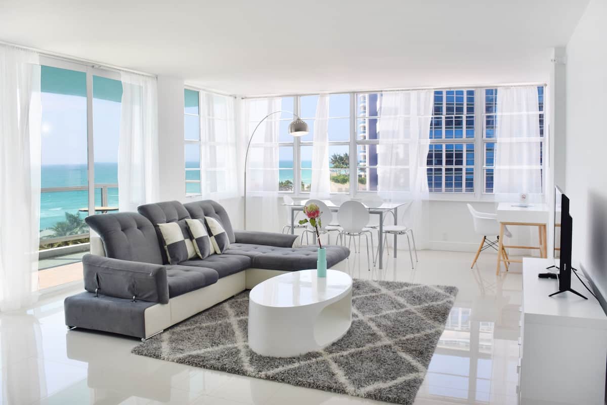 A modern living room with a grey sectional sofa, a white coffee table, and a view of the ocean through large windows.