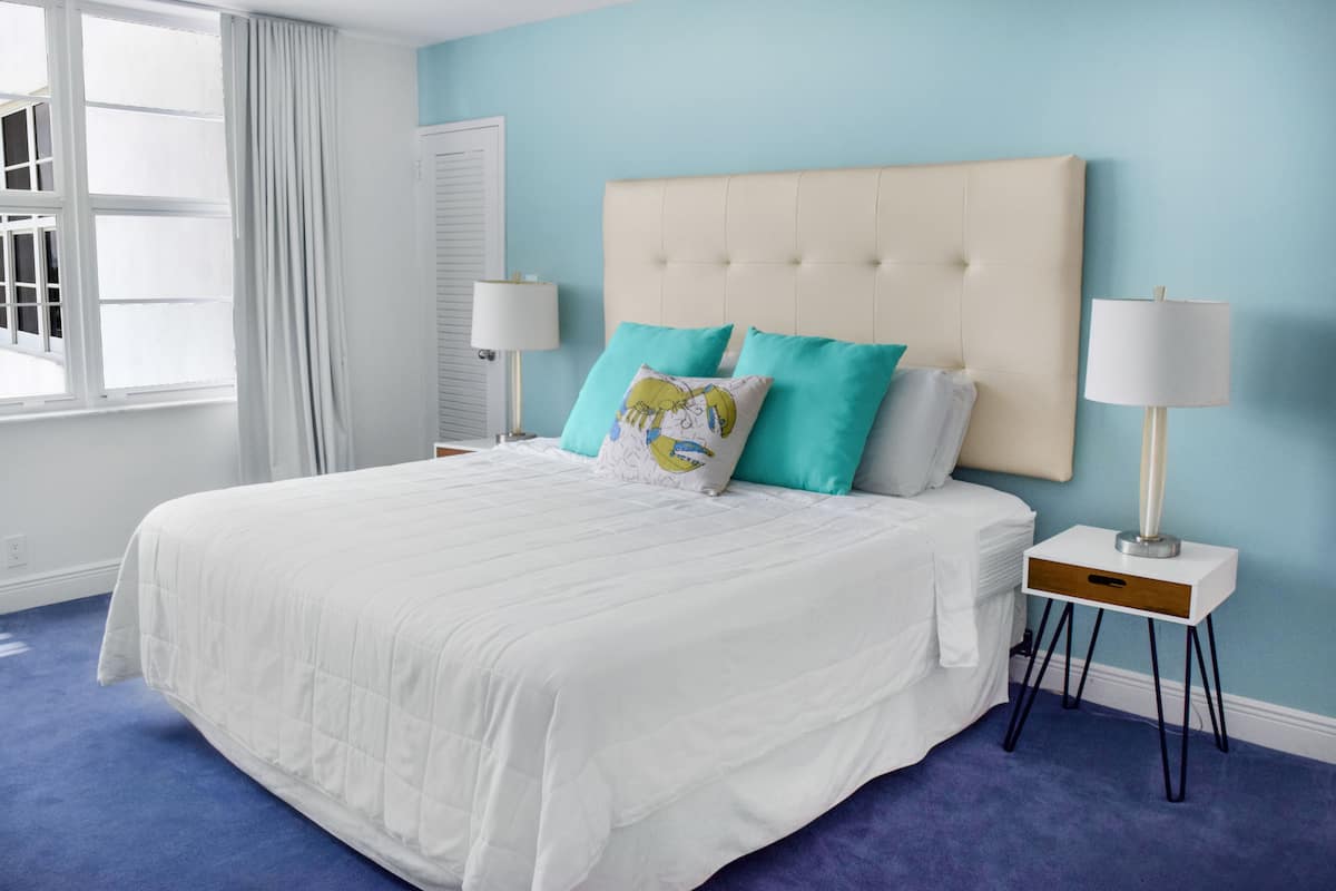 A bedroom with a large bed, two bedside tables with lamps, a window with curtains, and a light blue wall.