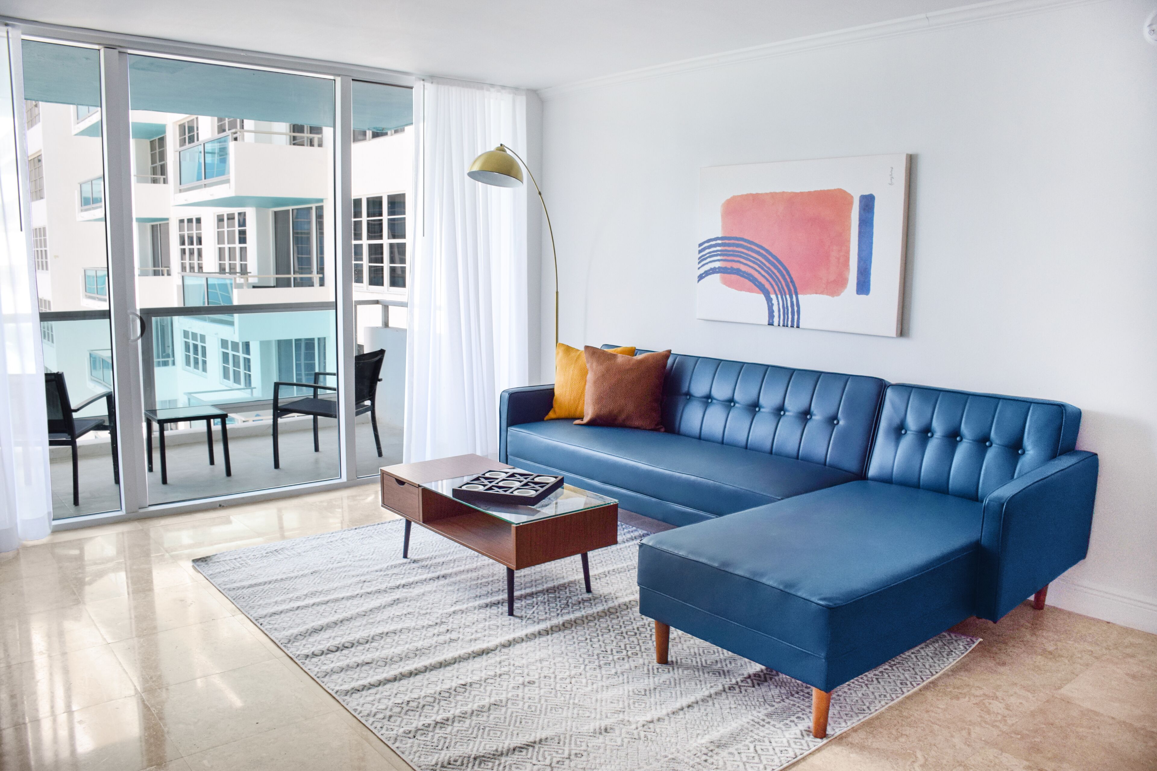 Ein modernes Wohnzimmer mit einem blauen Ecksofa, einem Glastisch und einem großen Fenster mit Blick auf ein Gebäude.