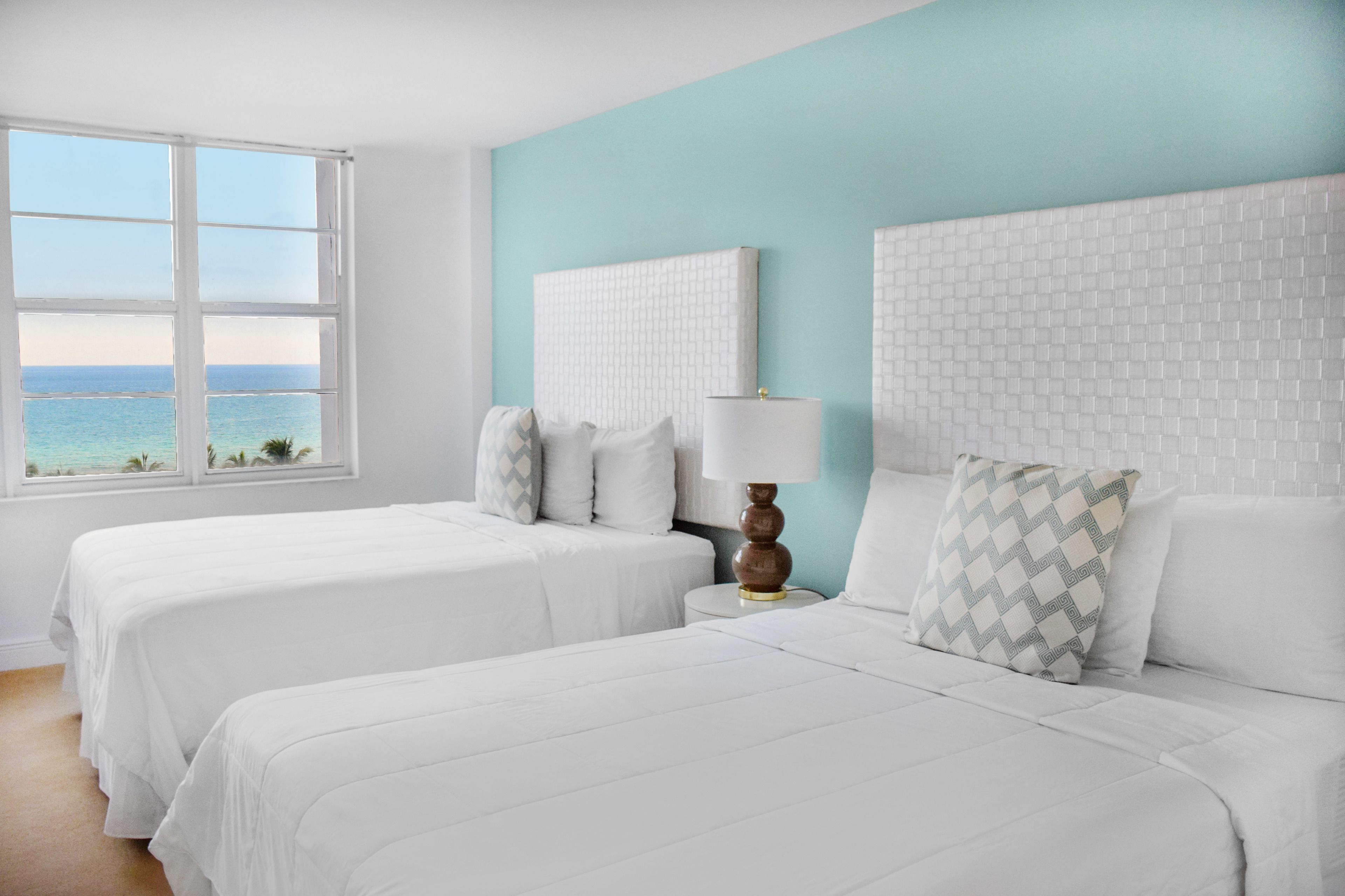 Une chambre d’hôtel avec deux lits, une grande fenêtre donnant sur l’océan et un mur recouvert de carreaux blancs.