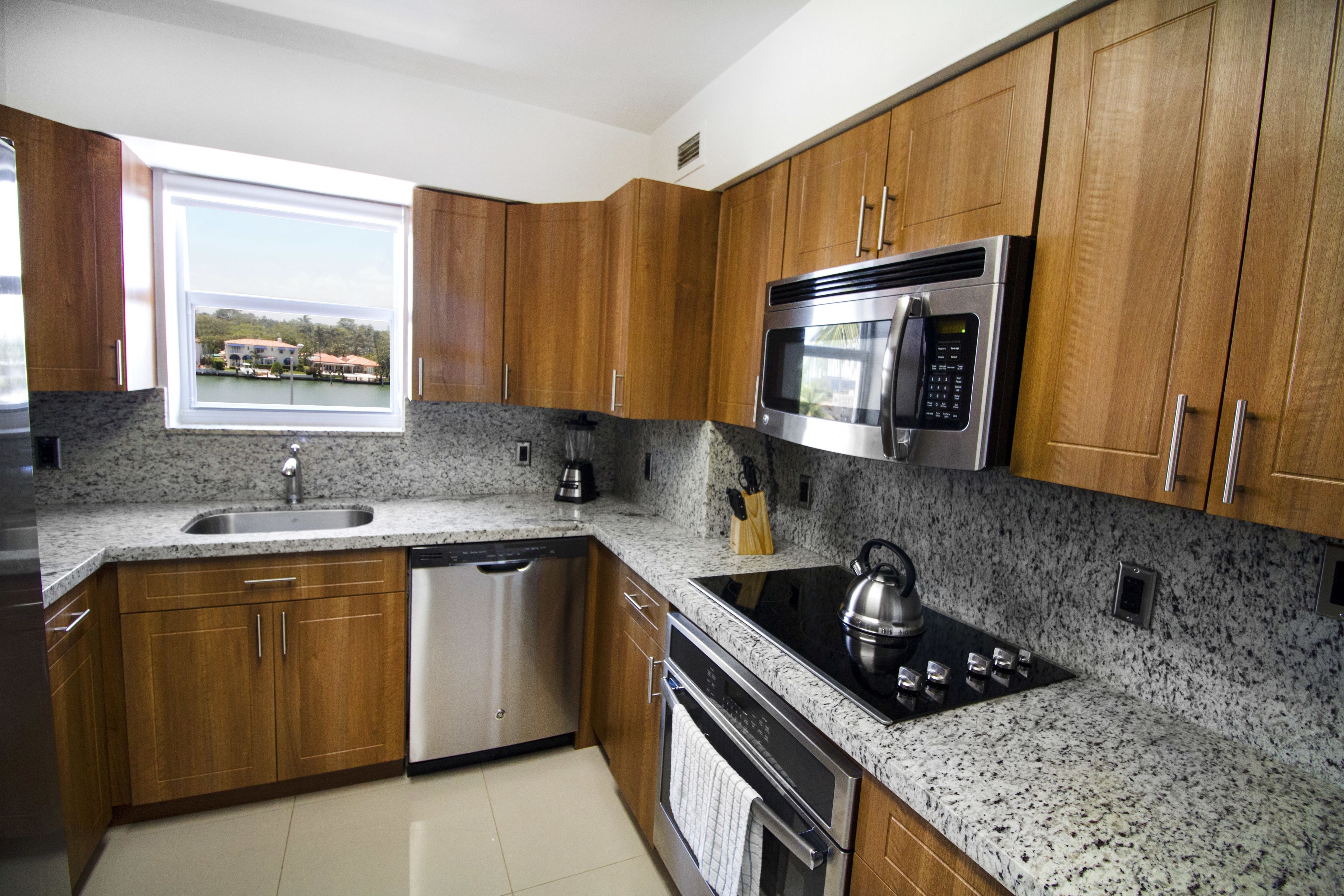 A modern kitchen with granite countertops, stainless steel appliances, wooden cabinets, and a window offering a view of a dock and boats.