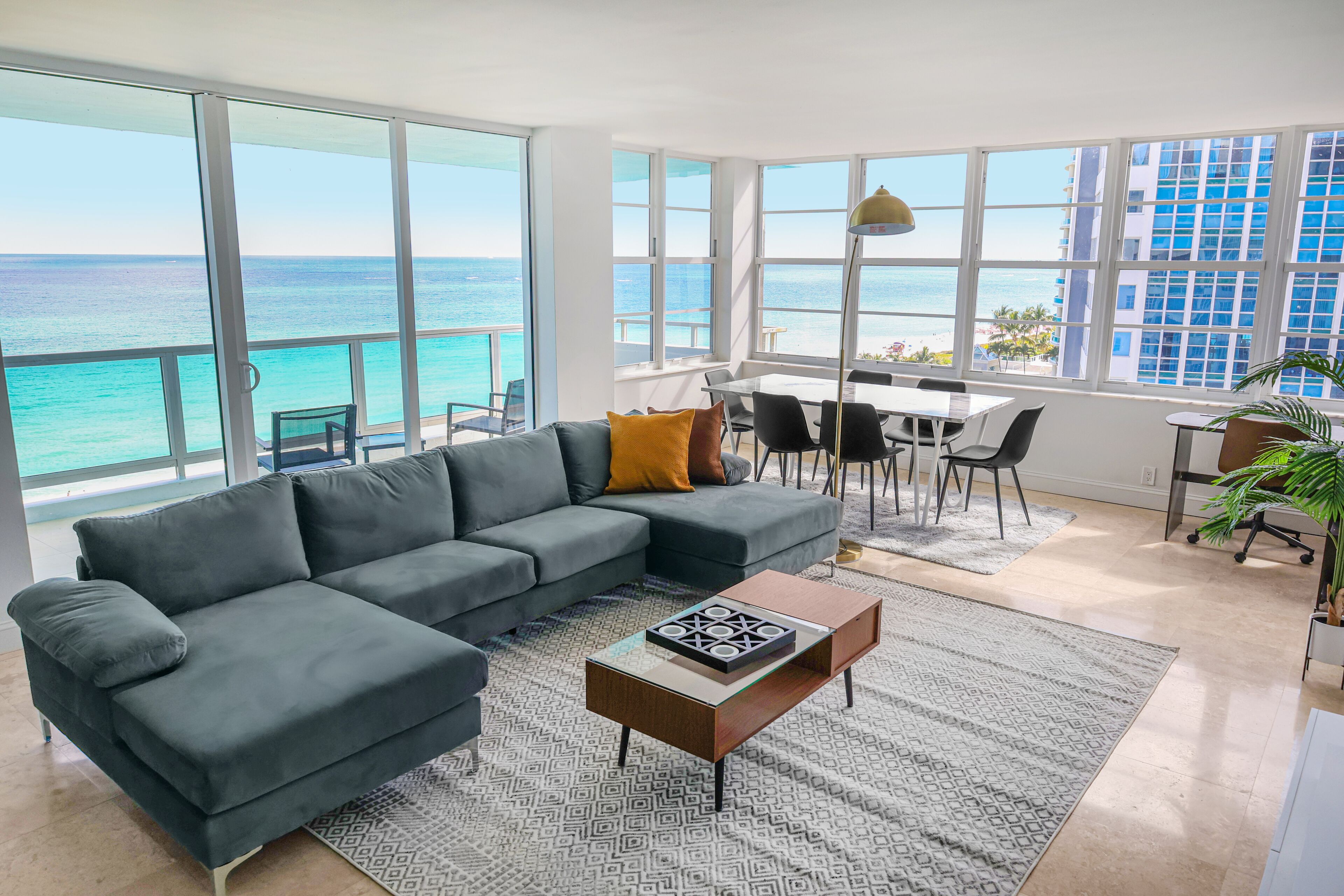 A modern living room with a large gray sectional sofa, a glass coffee table, and a view of the ocean through large windows.