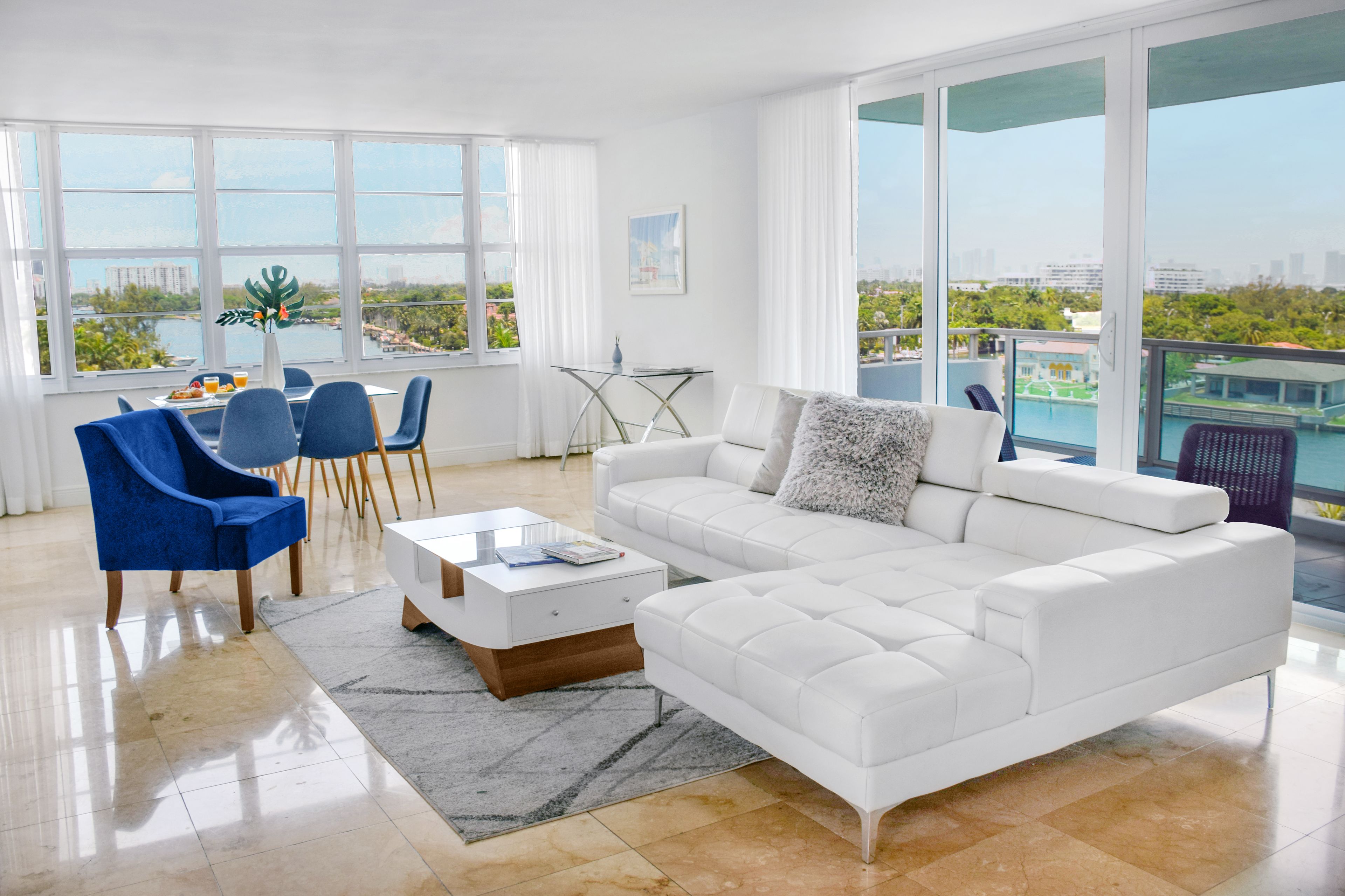 A modern living room with a white sofa, blue chairs, a glass coffee table, and a view of a cityscape through large windows.