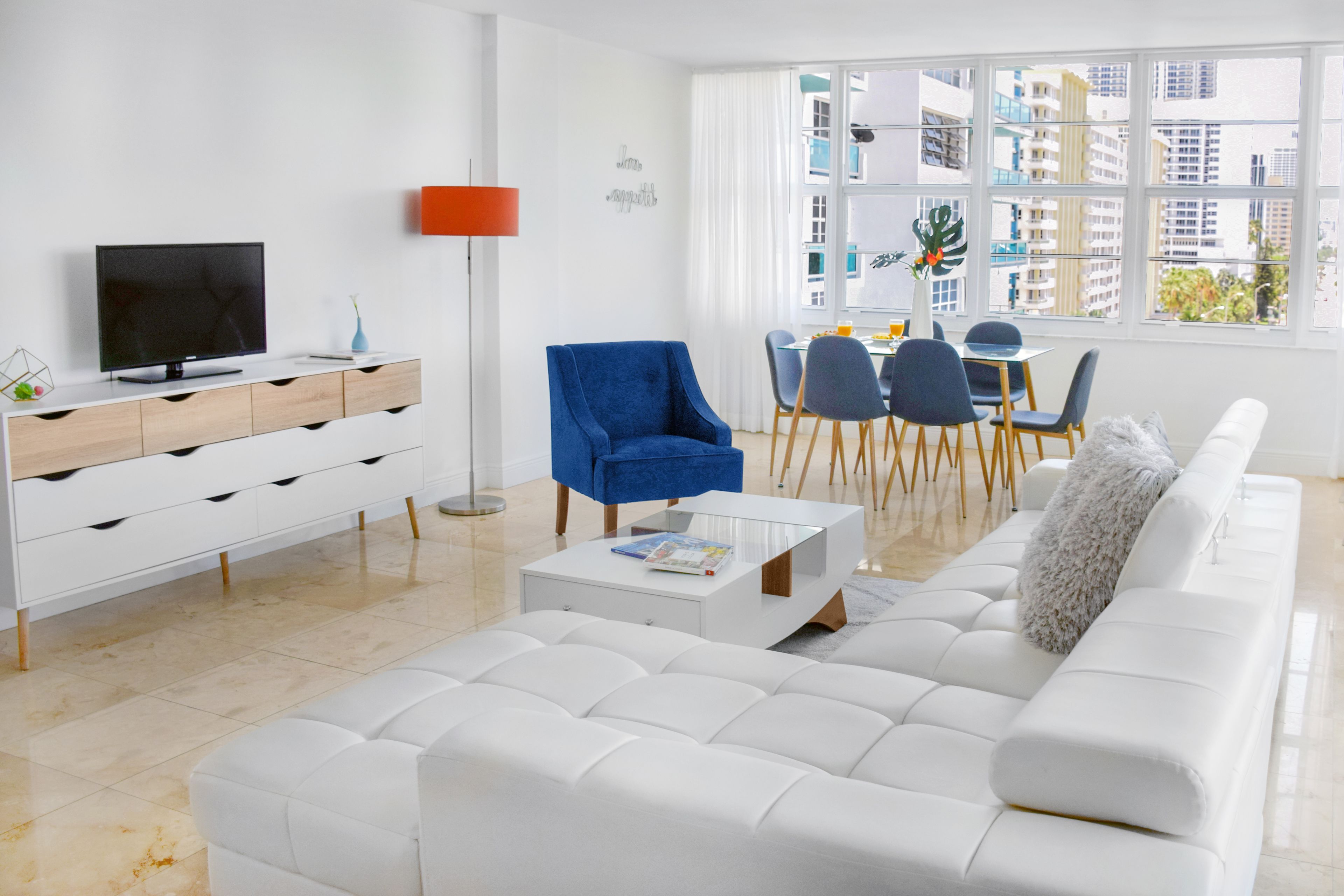 Een moderne woonkamer met een witte bank, een blauwe fauteuil, een tv op een witte dressoir, en een eethoek met blauwe stoelen en een tafel.