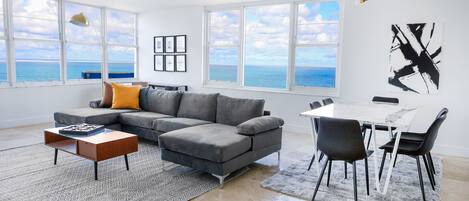 A modern living room with a gray sectional sofa, a small wooden coffee table, a dining table with chairs, and a view of the ocean through large windows.