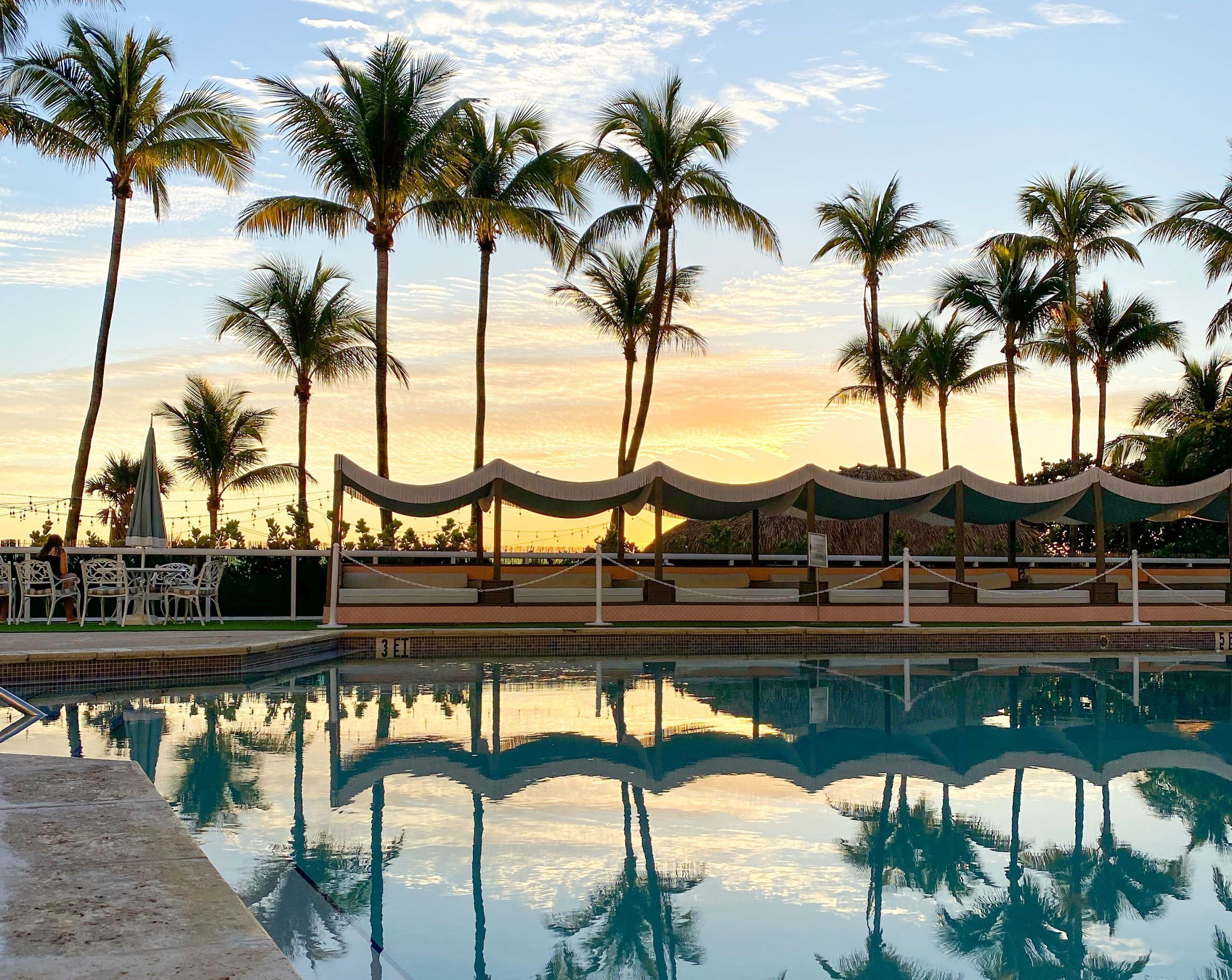 A pool area with lounge chairs and umbrellas, surrounded by palm trees and a sunset view.