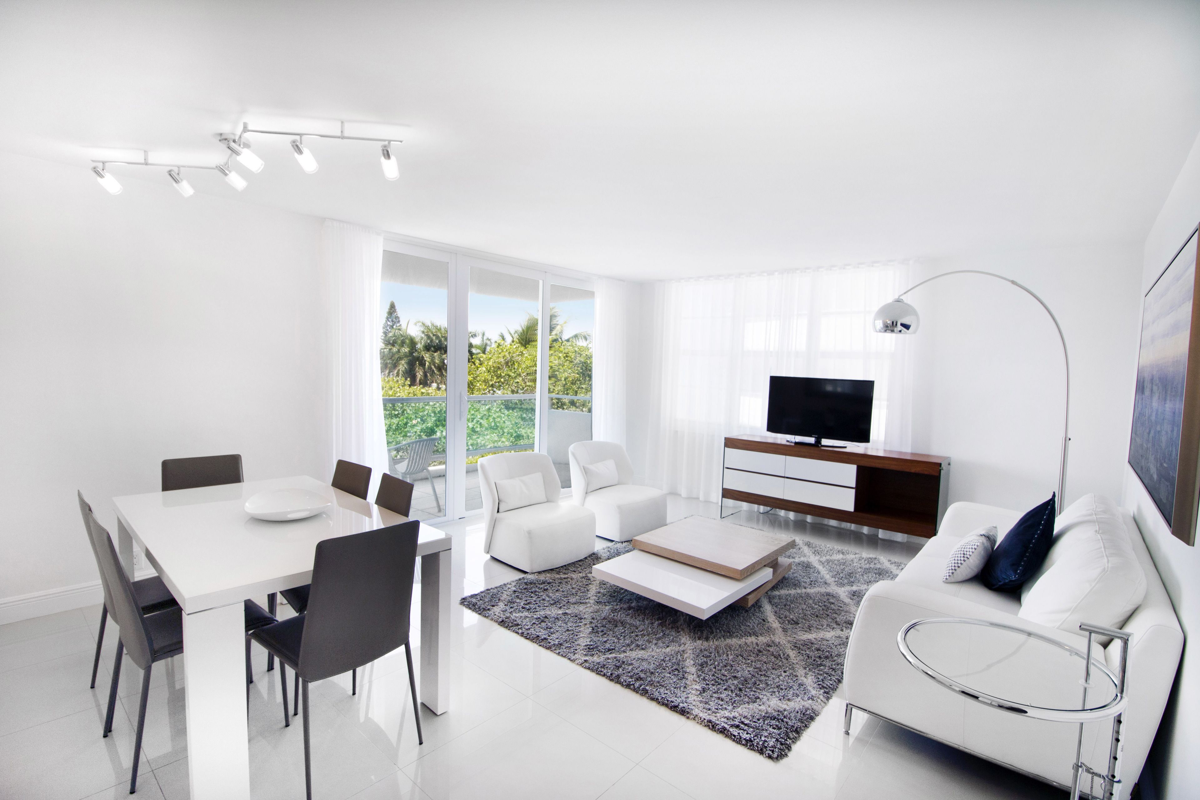 A modern living room with a dining area, white furniture, a flat-screen TV, and large windows.