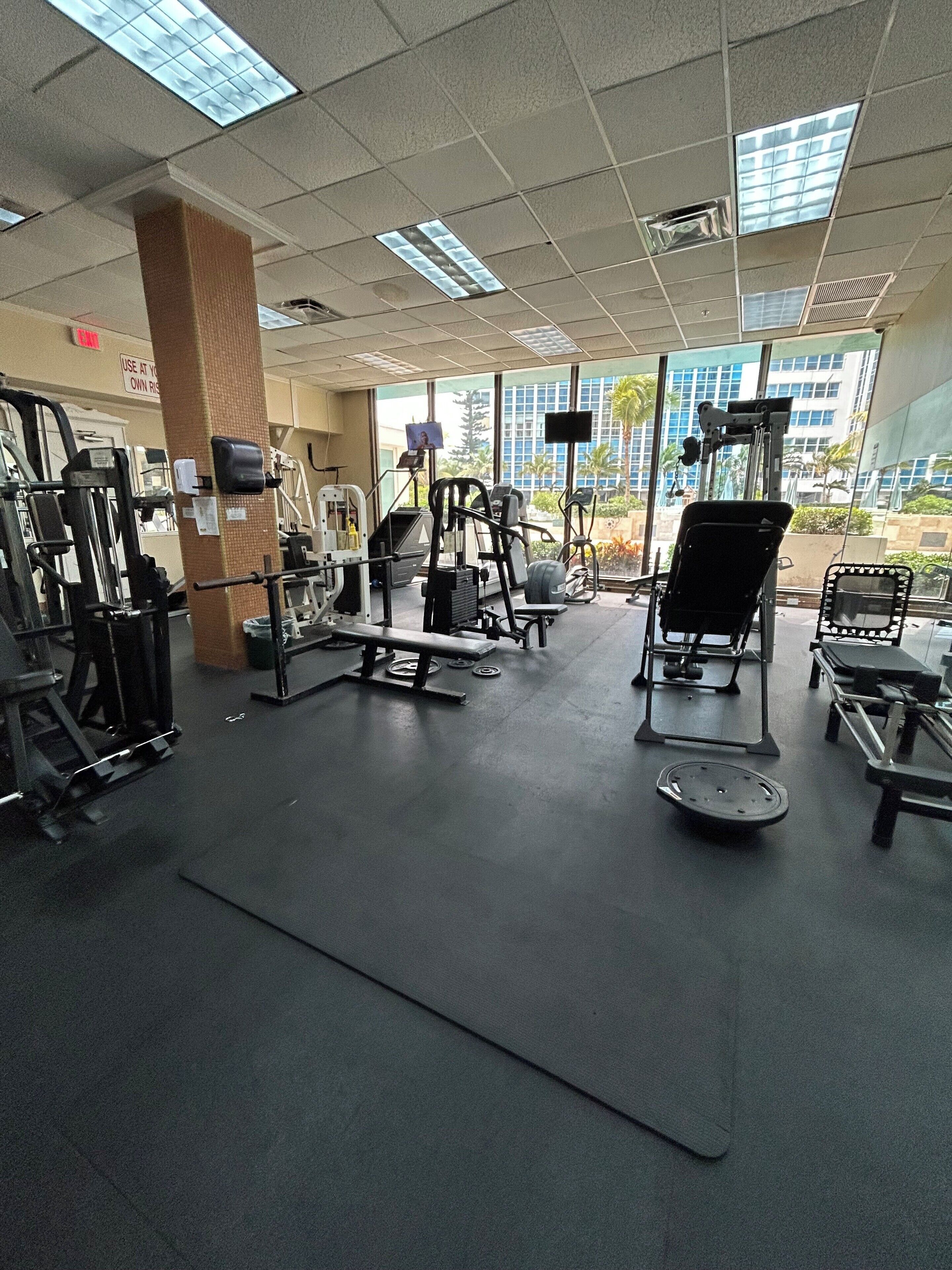 Un gimnasio con diverso equipo de ejercicio, ventanas amplias y vista a un edificio exterior.