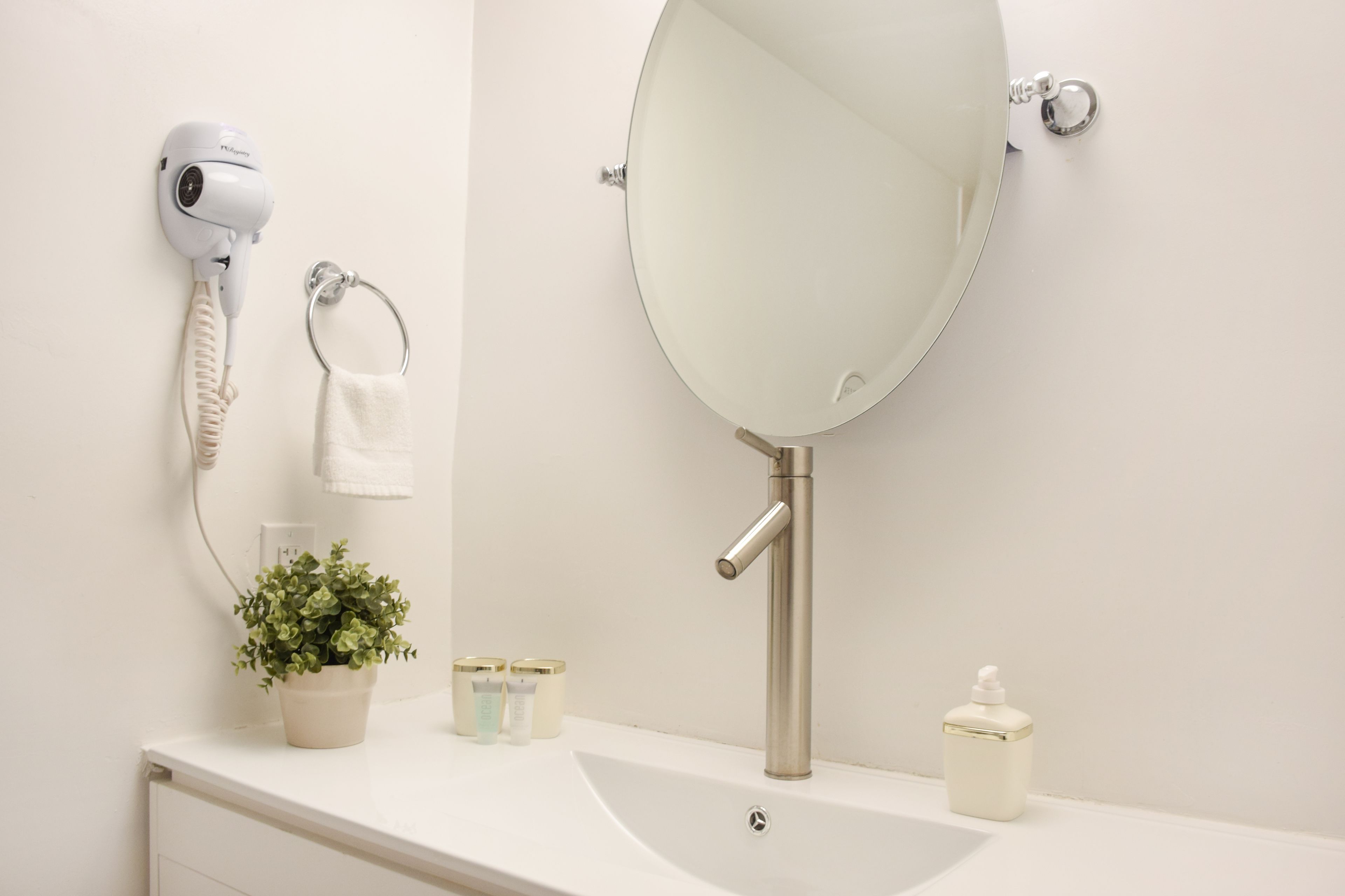 A bathroom with a white sink, a round mirror, a hairdryer, a towel ring, a potted plant, and two toiletry containers.