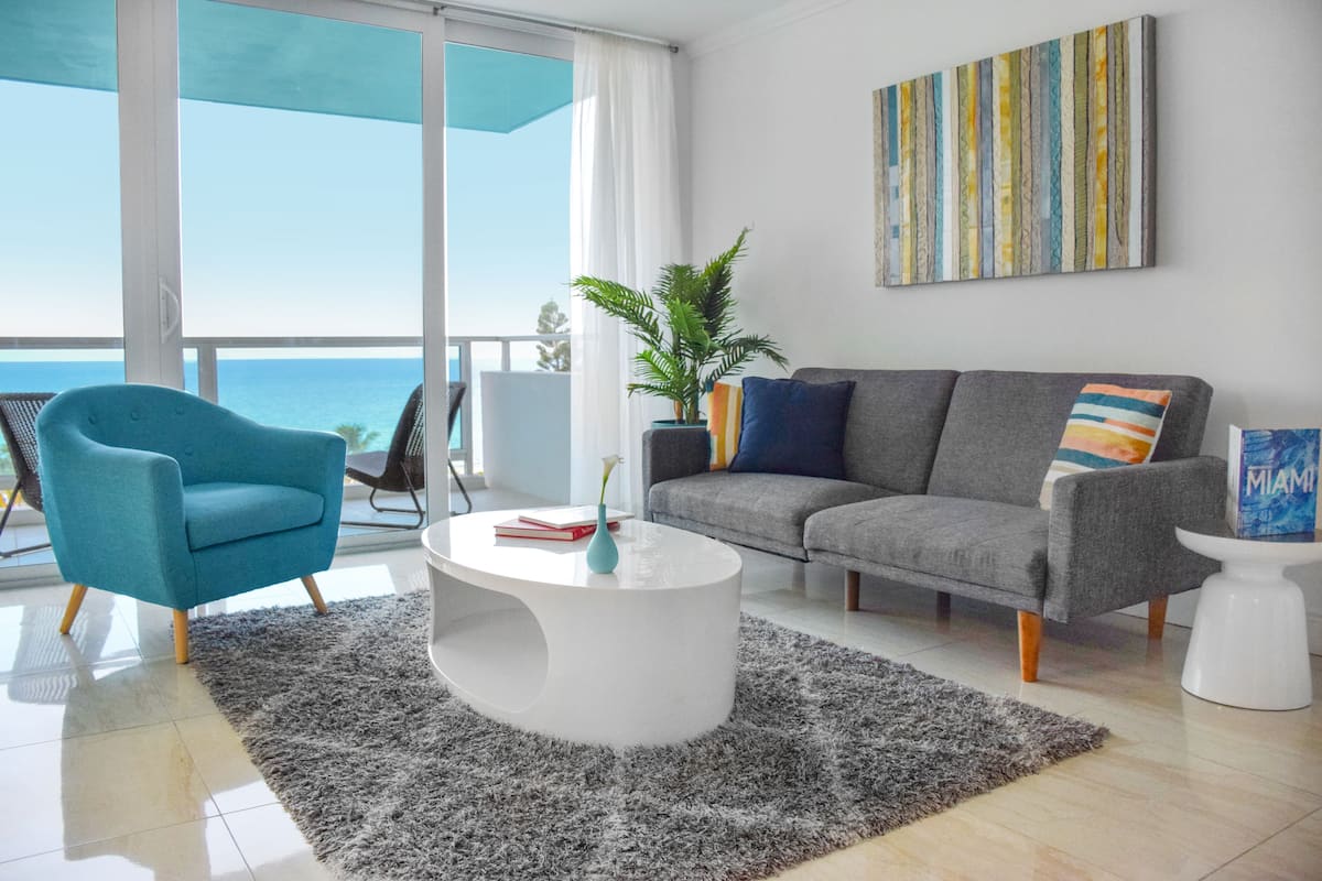 A modern living room with a grey sofa, a blue armchair, a white coffee table, and a view of the ocean.